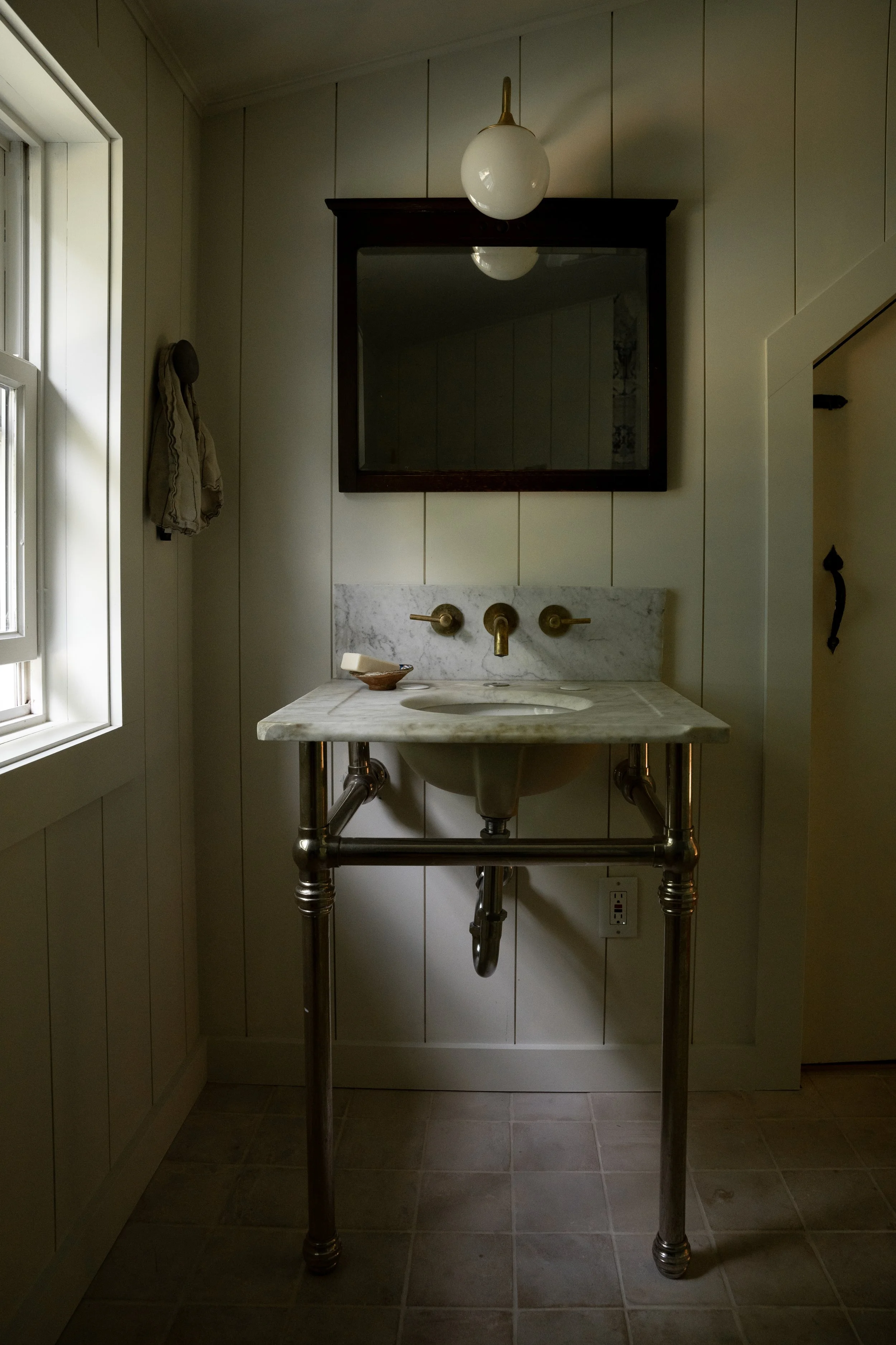 A bathroom with a marble sink and brass fixtures, a mirror above the sink, and a light fixture with a glass globe. There is a window on the left side and a towel hanging on a hook.