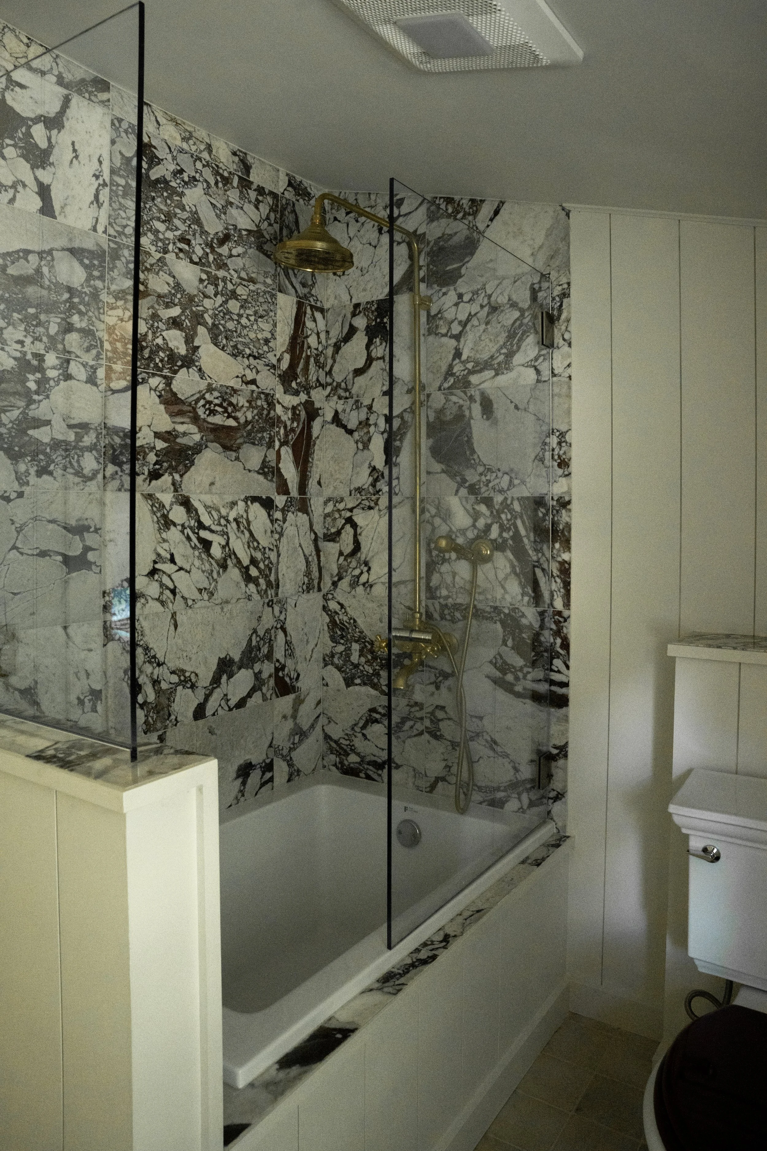 A marble-tiled shower with a brass rain showerhead and a handheld shower attached to the wall, with white cabinetry and a white toilet nearby in a modern bathroom.