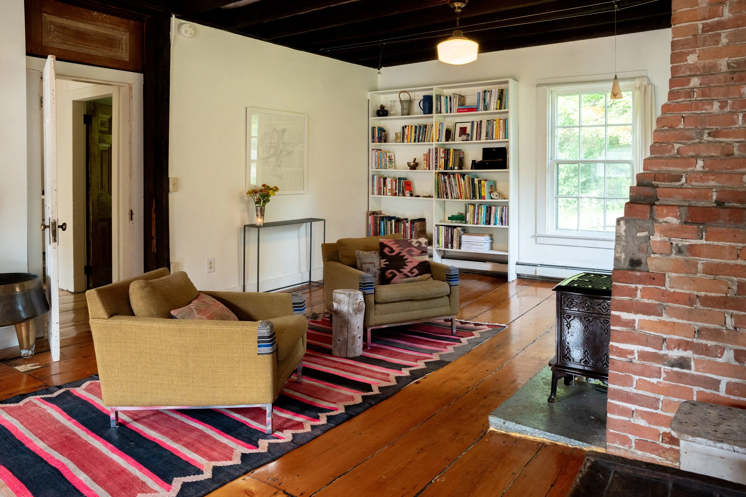 Living room with wooden floors, a brick fireplace, a white bookshelf filled with books, and seating including beige armchairs with pillows. There's a window showing green trees outside.