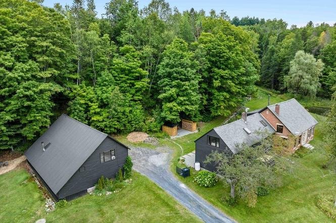 Aerial view of two houses surrounded by green trees and grass, with a gravel driveway connecting them.