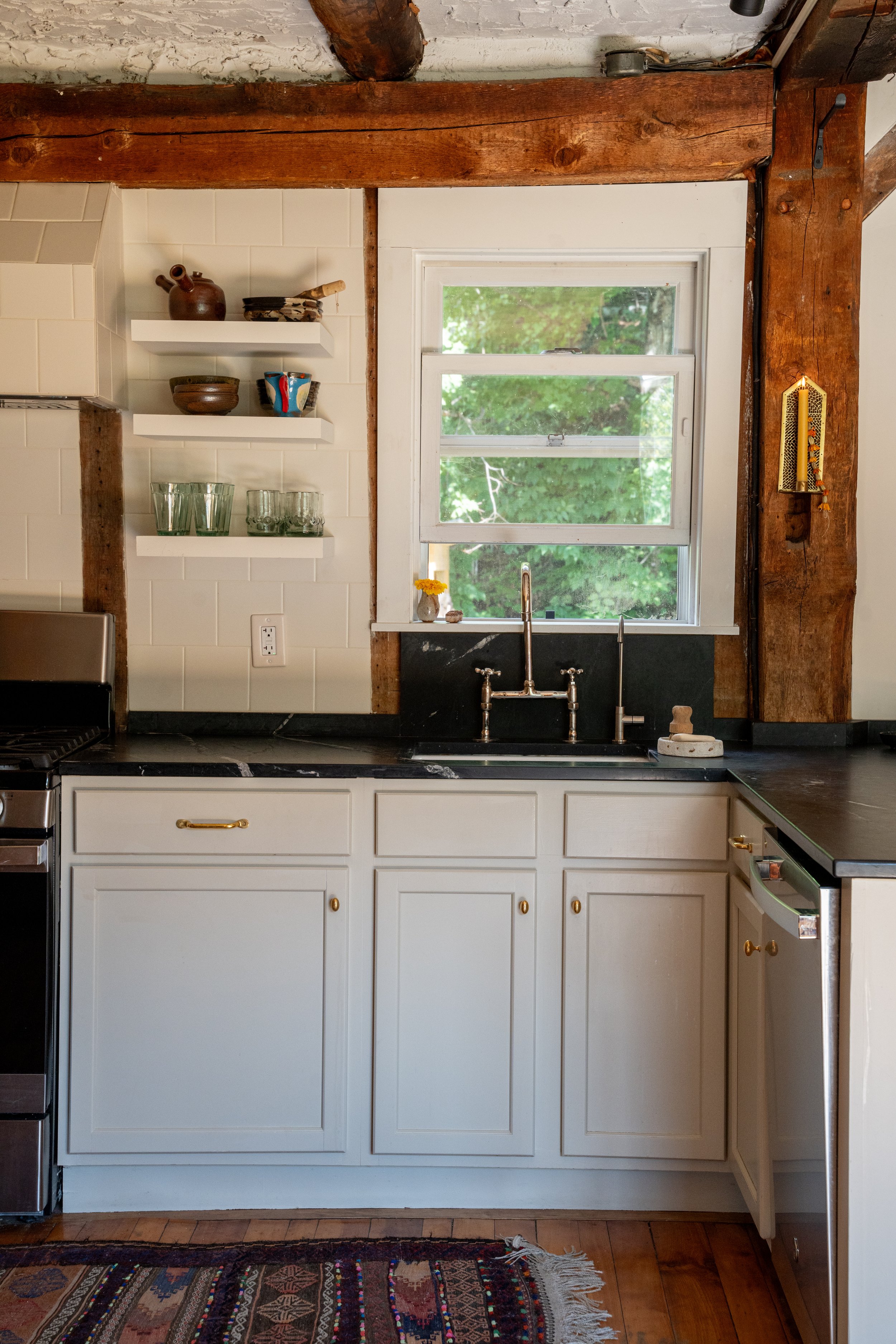 A kitchen area with a large window, white cabinetry with gold knobs, open shelves with bowls and cups, a black countertop, and wooden beams on the ceiling.