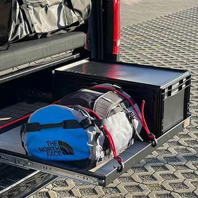 Ein Gepäckstück und eine große schwarze Box auf einer ausfahrbaren Plattform im Kofferraum eines Autos, mit einem Rollenkoffer und einem Rucksack darauf.
