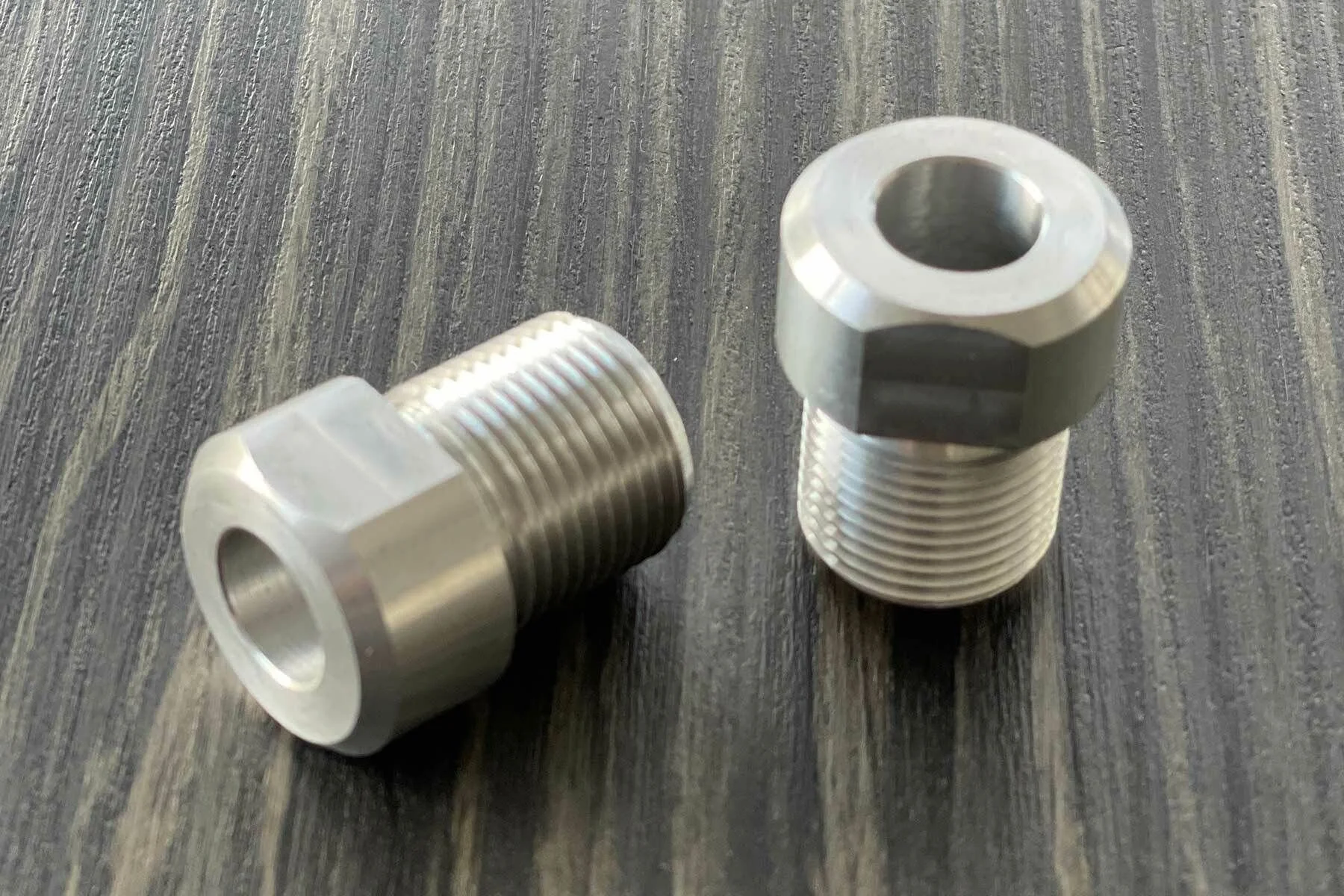 Zwei silberne Metallteile mit Außengewinde, die auf einer dunklen Holzoberfläche liegen.