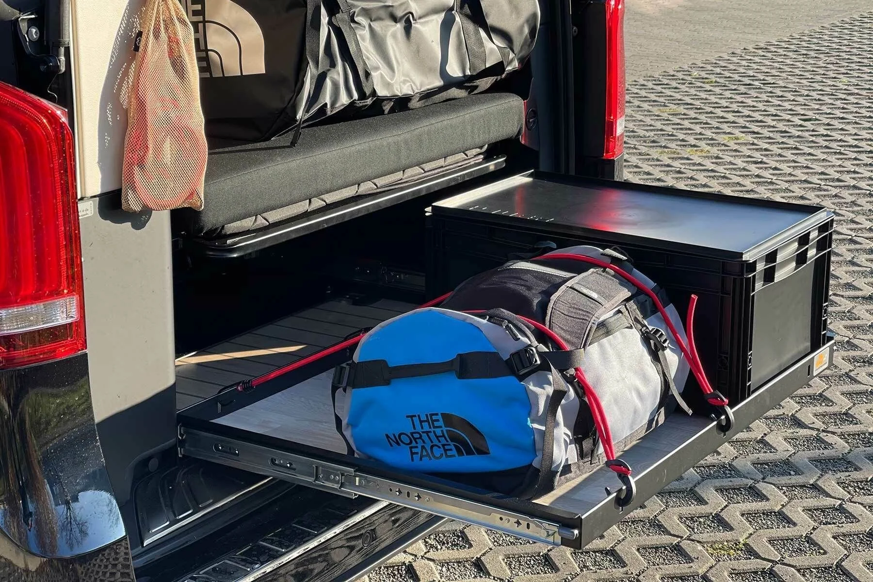 Offener Autokoffer mit Gepäck, darunter eine North Face Tasche, eine Kiste, ein Rucksack, eine Jacke, und weitere Gegenstände, auf einer gepflasterten Fläche.