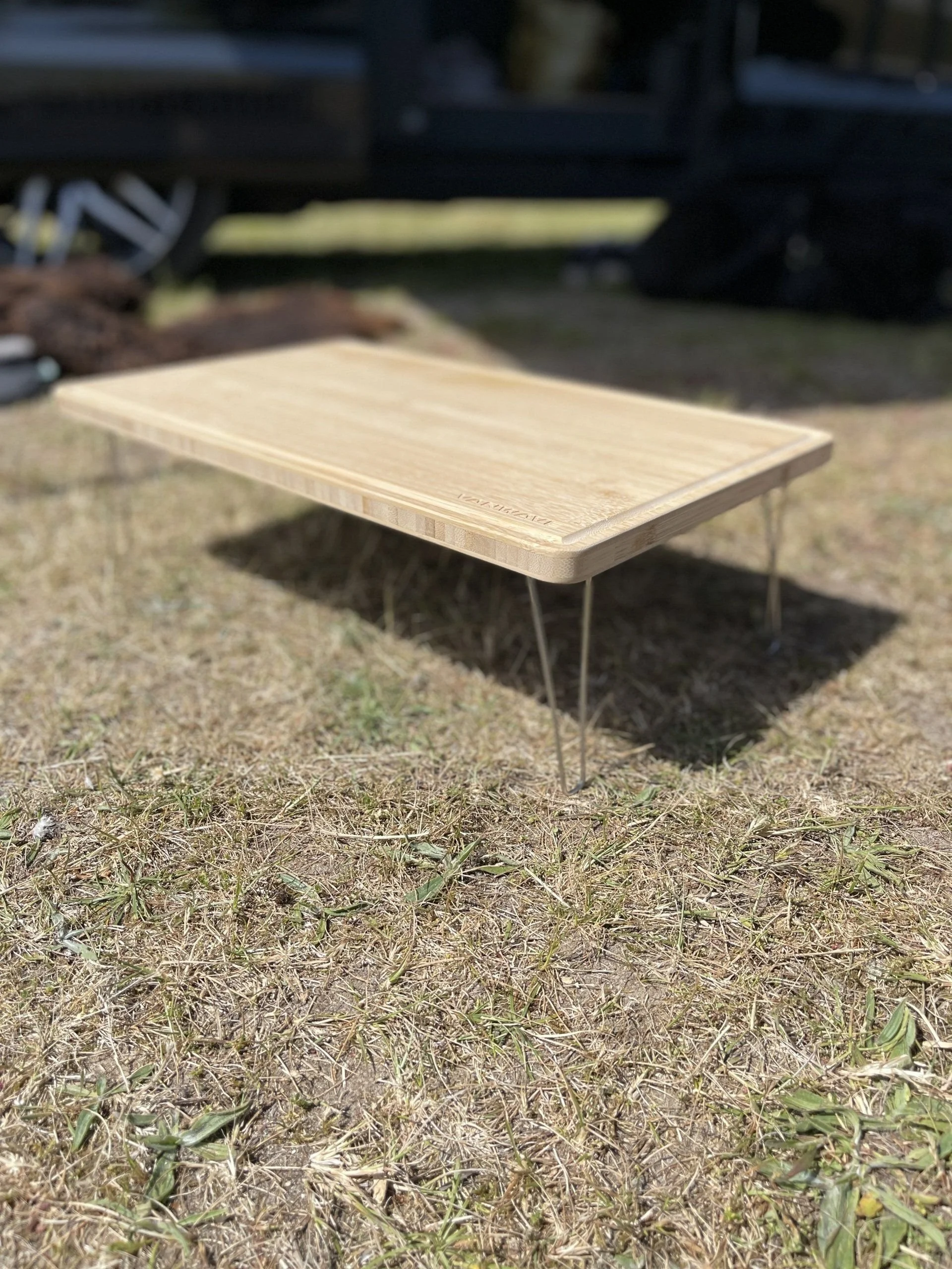 Ein Holztisch mit Metallbeinen steht auf trockenem Gras im Freien, im Hintergrund eine dunkle Karosserie eines Fahrzeugs.