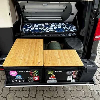 Zwei Holzplatten, die als Tische auf einer unteren Lagerfläche in einem Fahrzeug stehen, mit einer aufgeschlagenen oberen Ladefläche im Hintergrund, die einige Taschen und Gegenstände enthält.