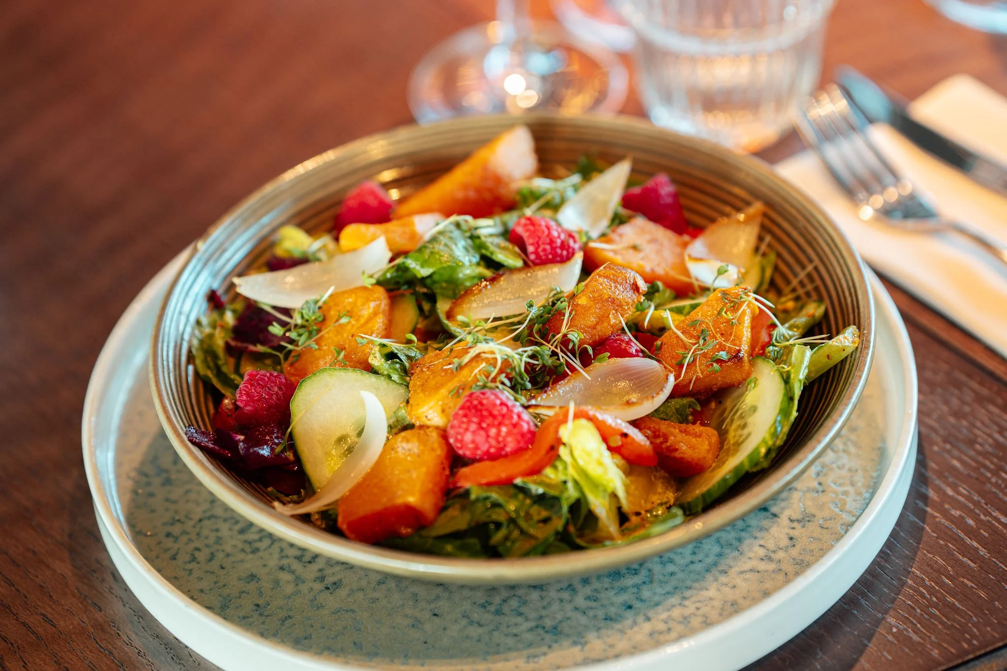 En bol med salat med røde bær, agurk, salatblader, og annet grønt og orange. På en trebord med glass vann og bestikk rundt.