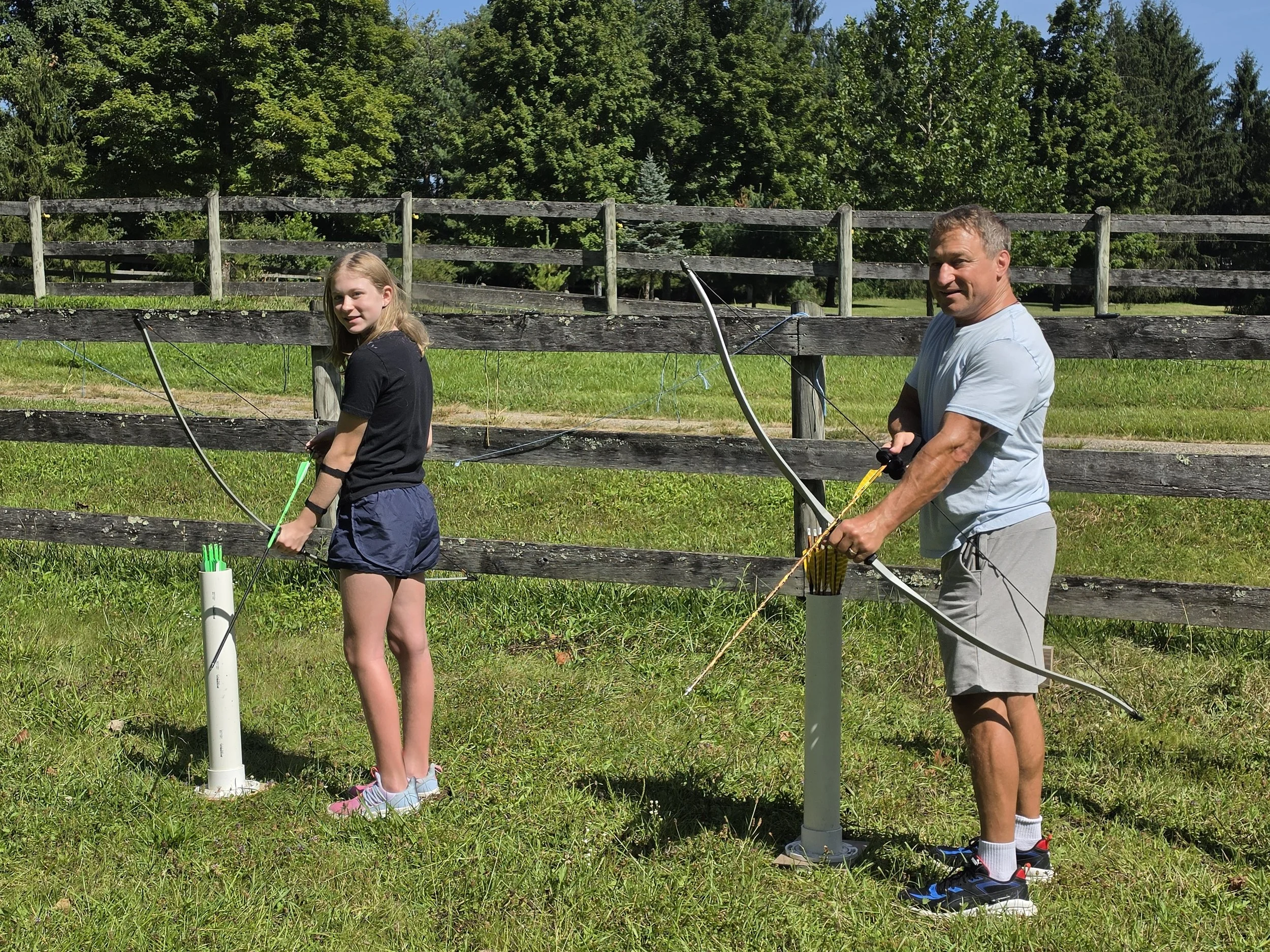 A young girl and an older man are outside behind a wooden fence, aiming bows and arrows at a target. The girl has blonde hair, is wearing a black t-shirt and blue shorts. The man has short gray hair, is wearing a light gray t-shirt, light gray shorts, and sneakers. There is a grassy area with trees and blue sky in the background.