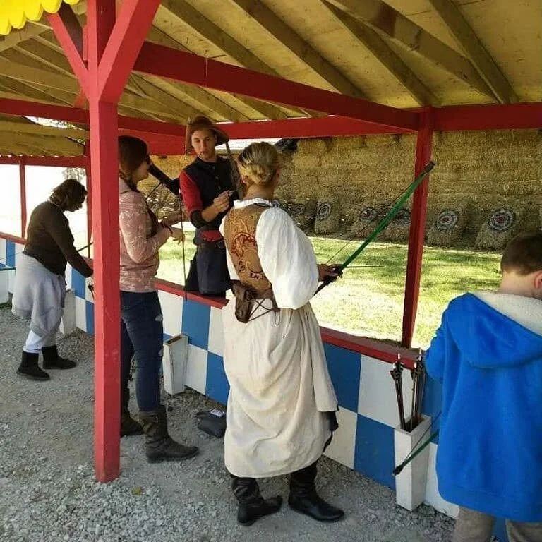 People at an outdoor archery range, some aiming with bows and arrows, some supporting or observing. The setting has a wooden roof, stone wall, and target boards in the background.