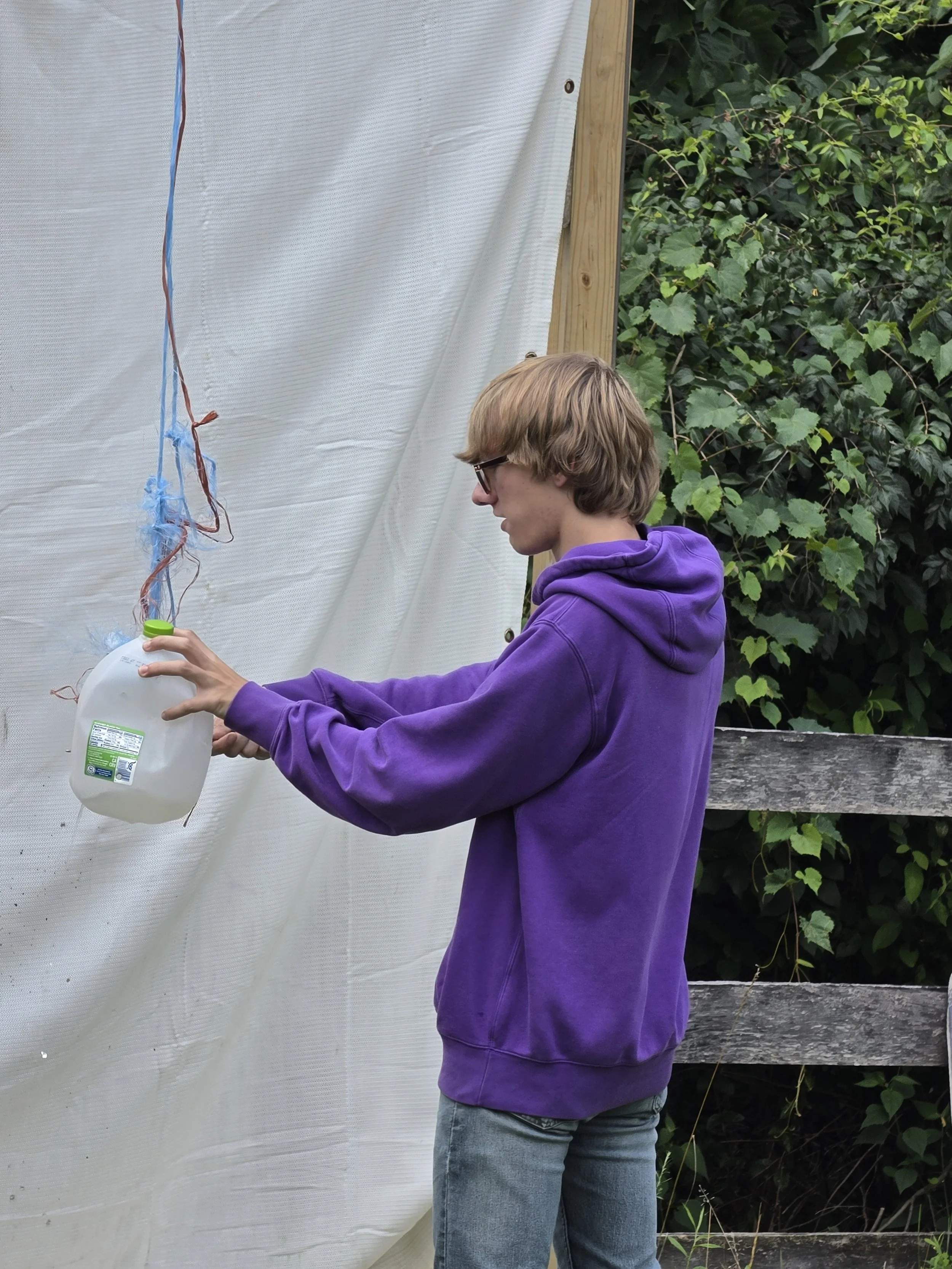 A young person with blonde hair, glasses, a purple hoodie, and jeans, filling a plastic jug with liquid from a container outdoors, standing in front of a white plastic sheeting and green foliage.