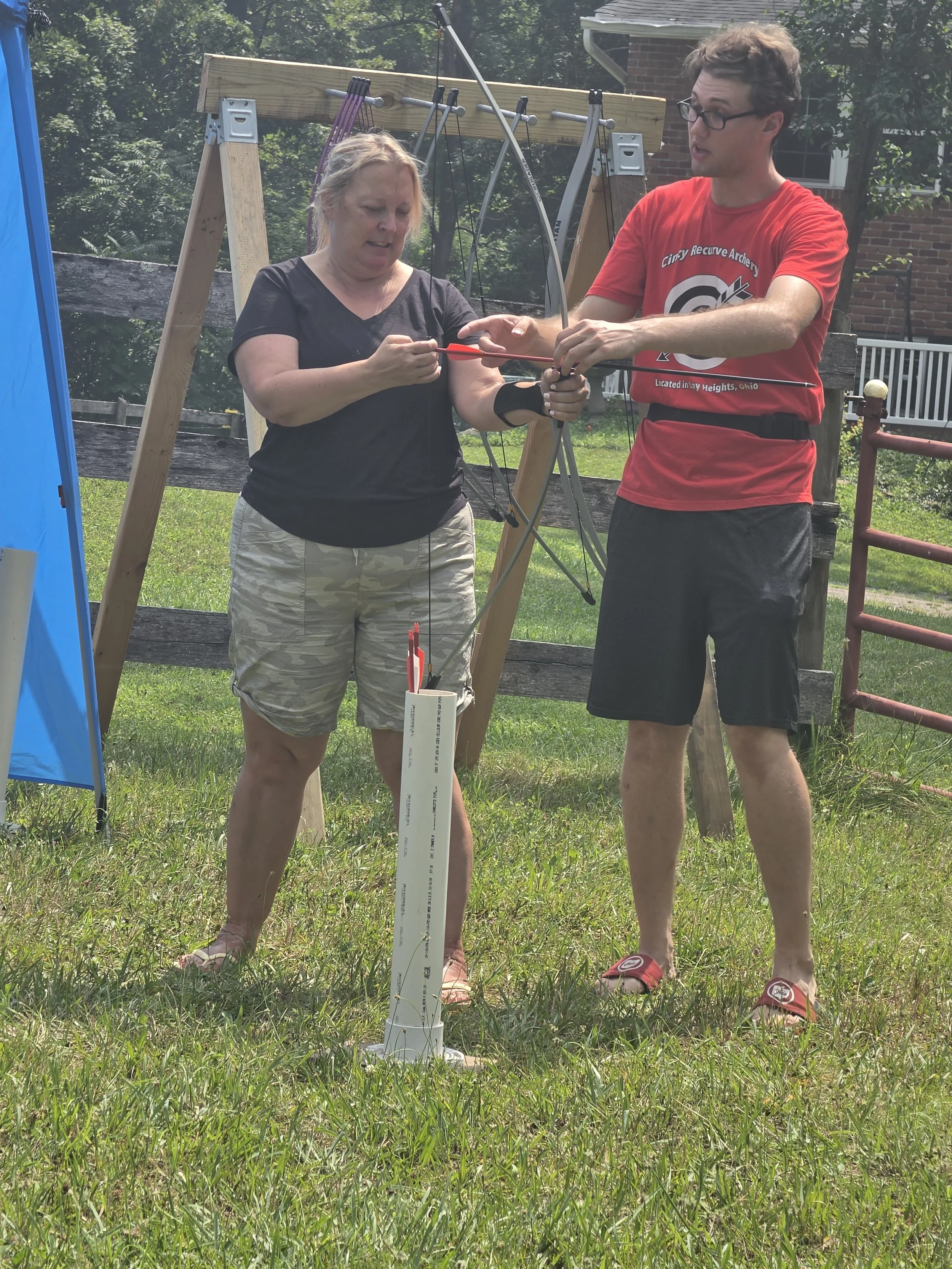 A man and woman outdoors near archery equipment, practicing shooting with a bow and arrow. The woman is holding an arrow, and the man is assisting her. There is a wooden frame with bows hanging on it, and a target is partially visible on the left.
