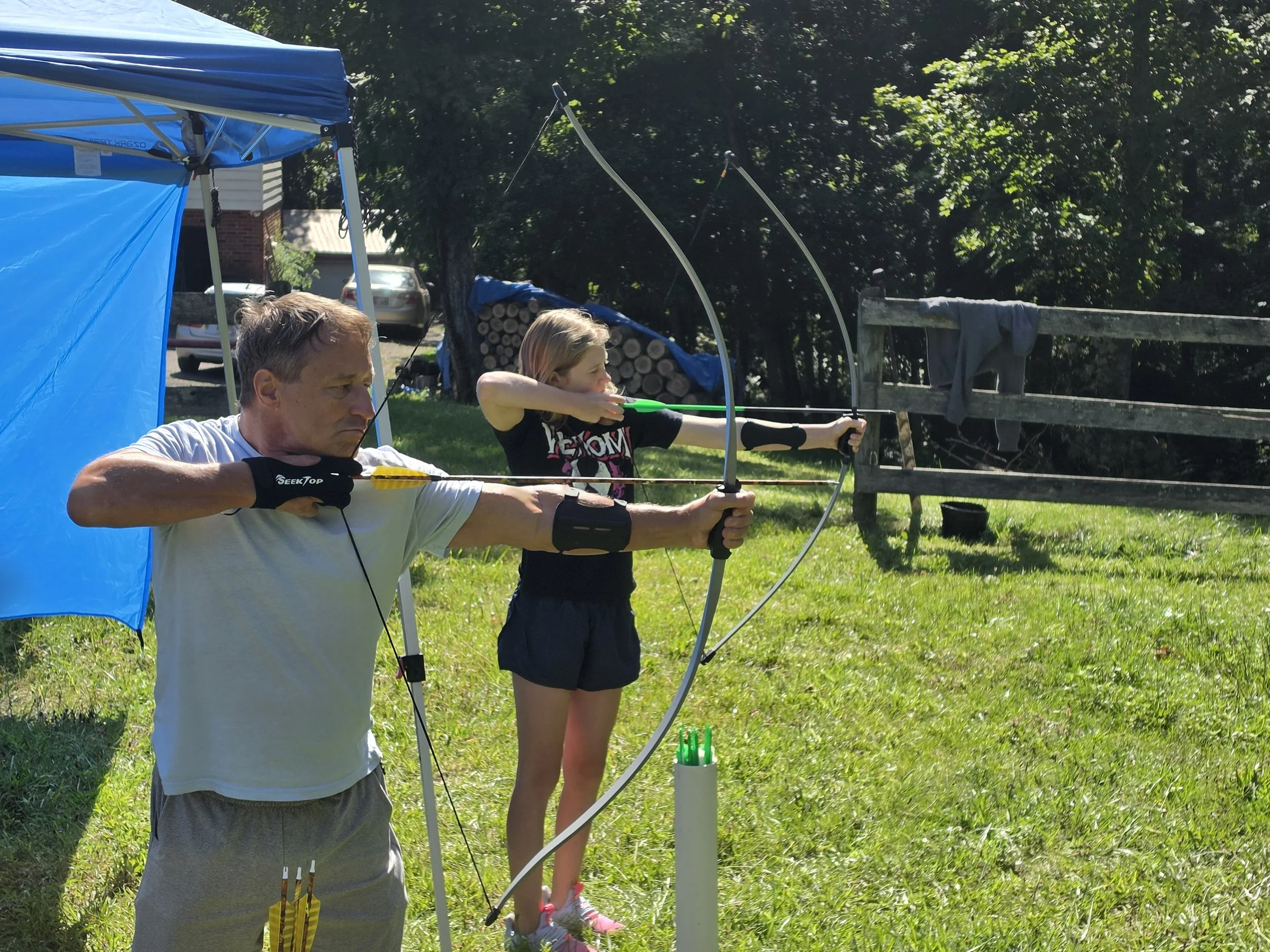 A man and a girl practicing archery outdoors on a grassy area, with trees in the background. The man is aiming a bow and arrow while the girl behind him is also shooting. There is a blue tent and logs nearby.
