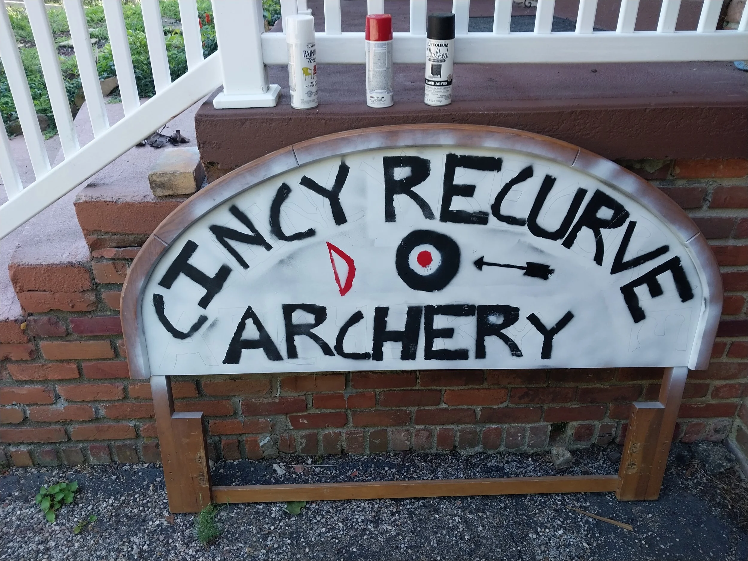 Close-up of an archery target sign with black text and a red arrow, leaning against a brick and wood structure, with cans of spray paint on top.