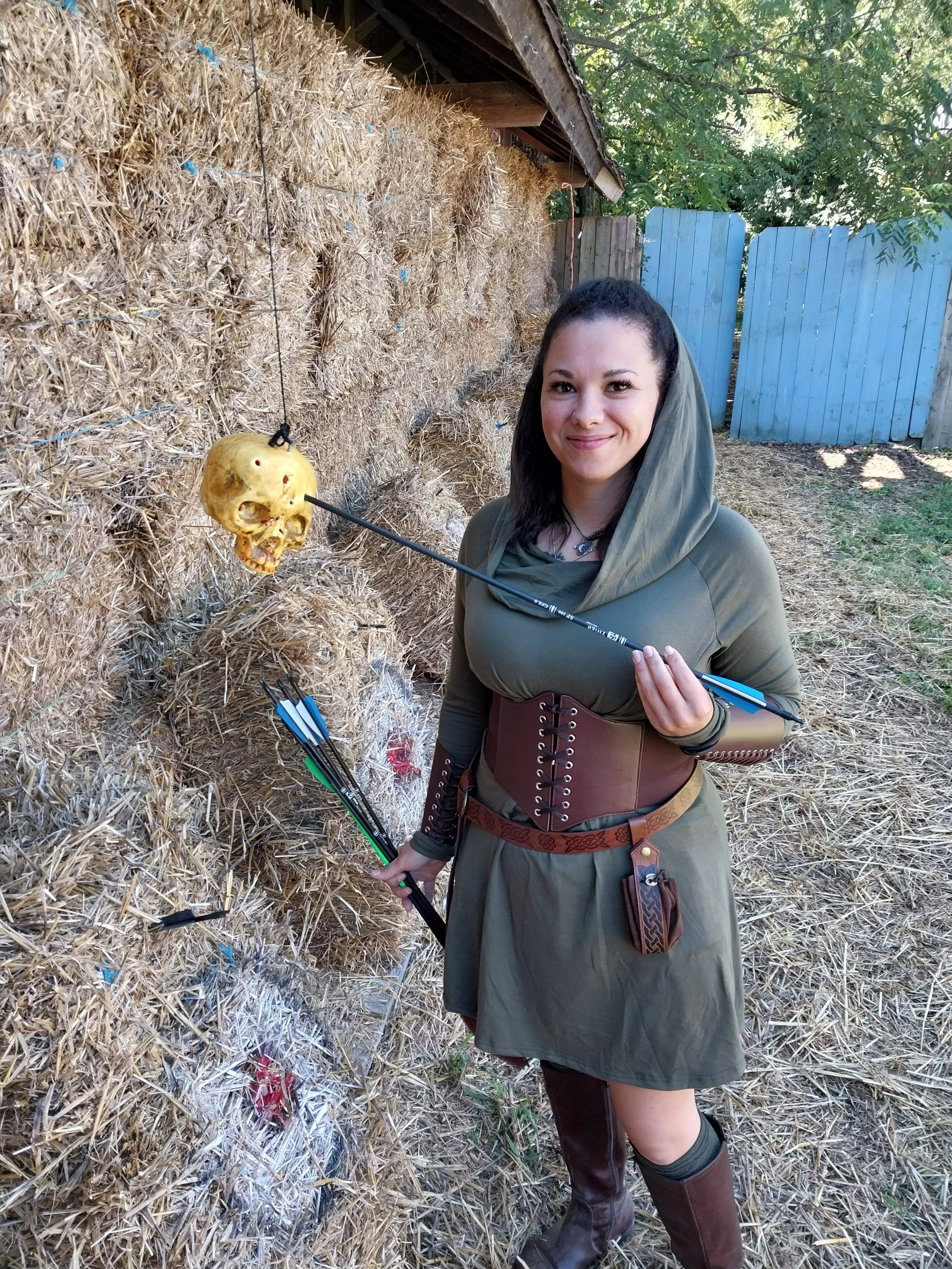 A woman dressed as a medieval or fantasy character standing outdoors next to a haystack with a blue wooden fence and trees in the background. She is holding a bow and has a skull ornament on a string around her neck.