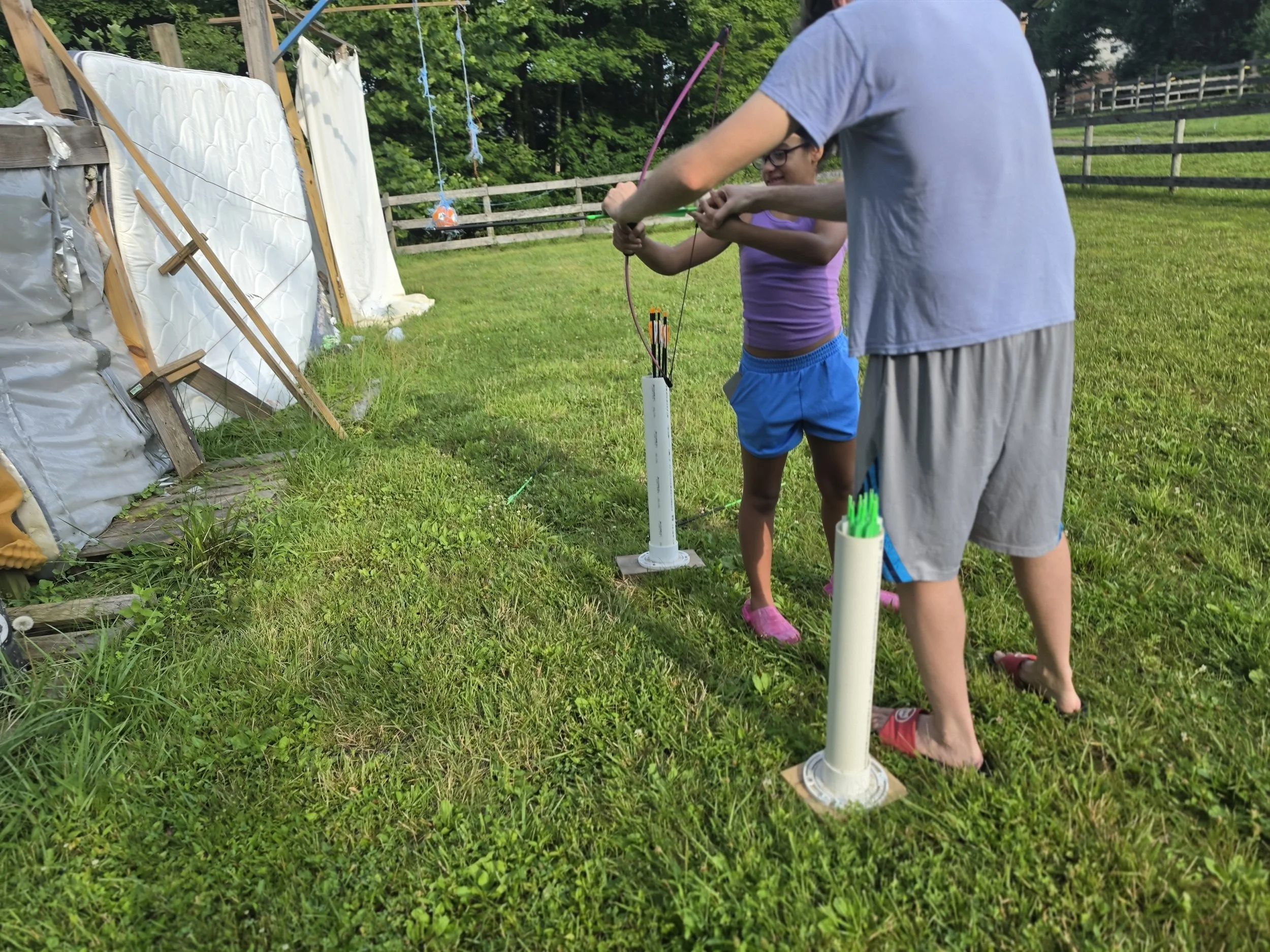 A girl and a man are outdoors, preparing to shoot arrows at a makeshift archery target made of mattress and wood. The girl is smiling, wearing glasses, a purple shirt, and blue shorts. The man is wearing a gray shirt and shorts. There are green fields and a wooden fence in the background.