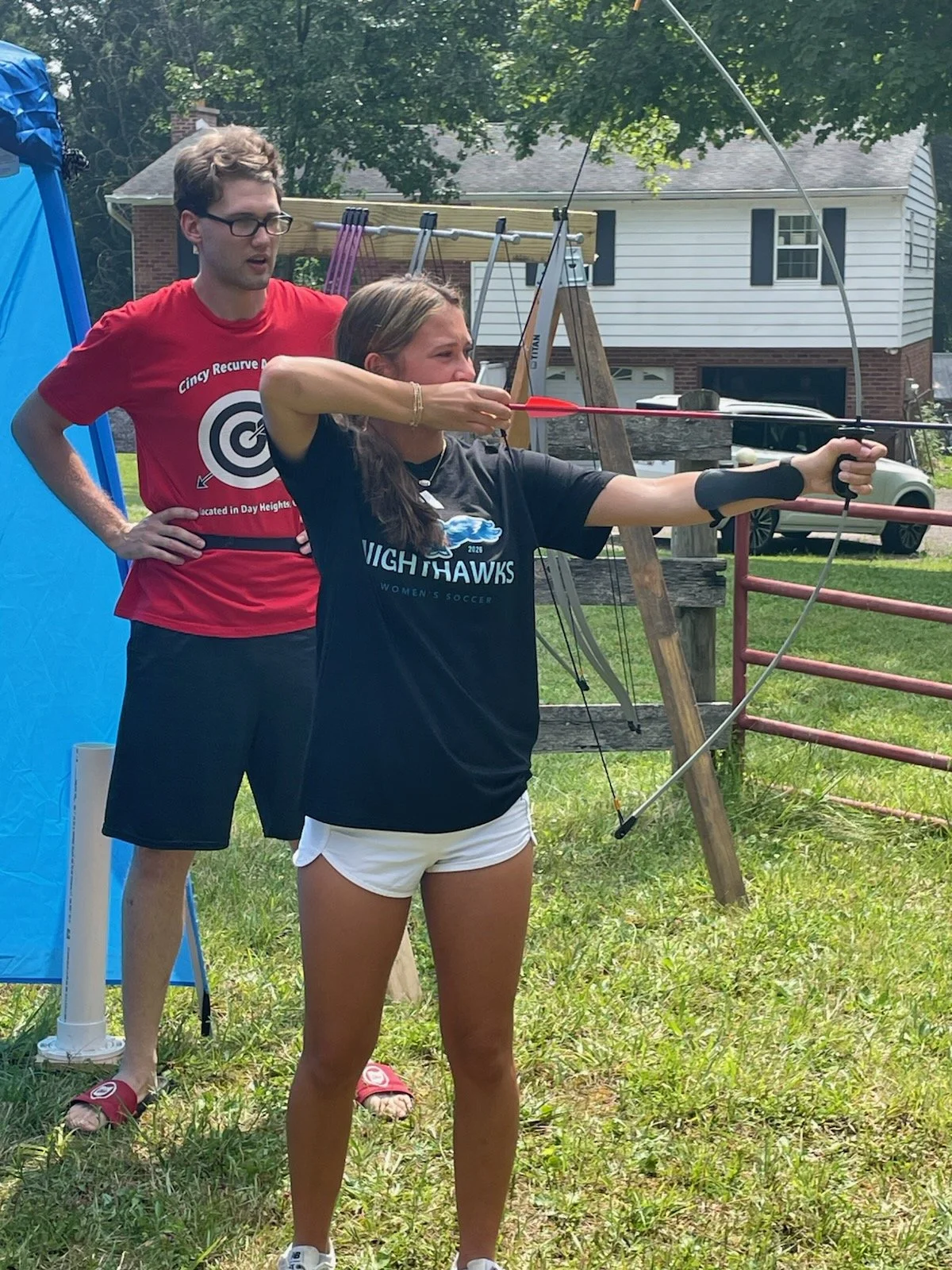 A young woman is practicing archery outdoors, aiming her bow and arrow, with a male instructor standing behind her observing, both on a grassy area with trees and residential houses in the background.
