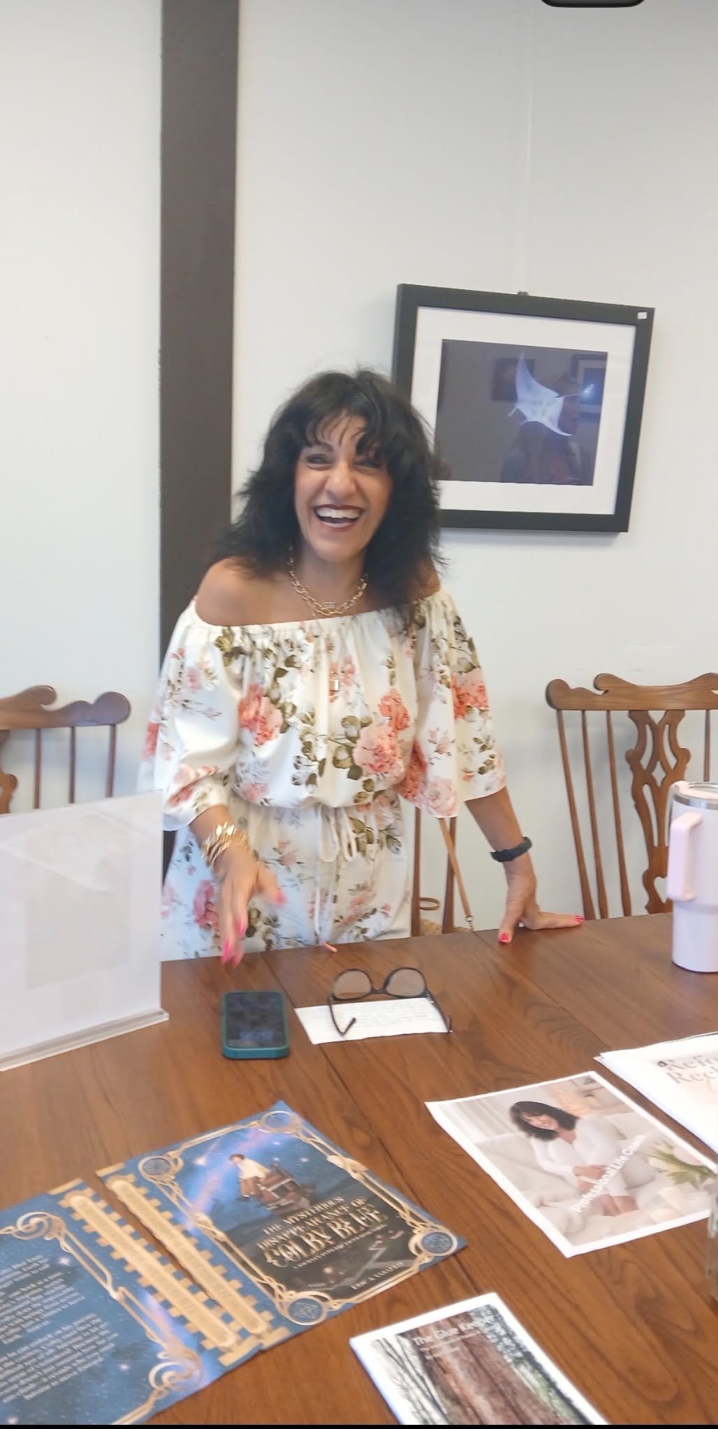 A woman in a floral dress is smiling and leaning on a wooden dining table with brochures, a smartphone, a pair of glasses, and papers on it. She is standing in a brightly lit room with wooden chairs and framed artwork on the wall.