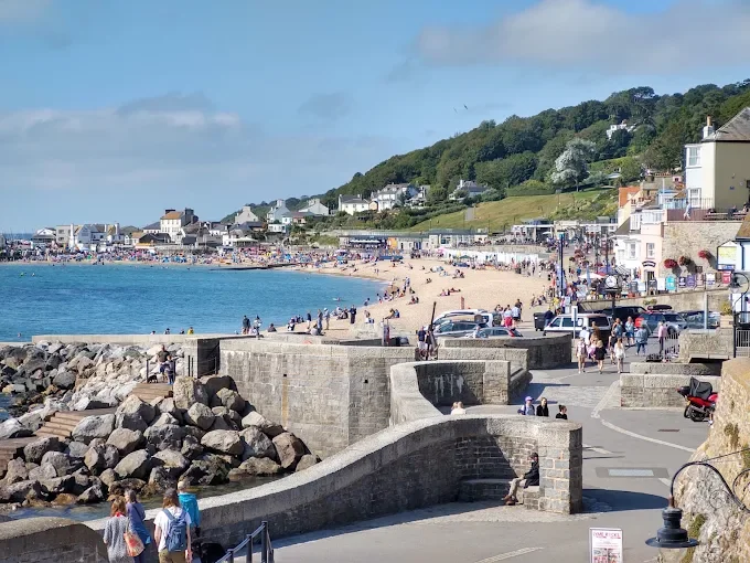 A busy seaside town with a sandy beach, people walking along the promenade, colorful buildings, and green hills in the background.
