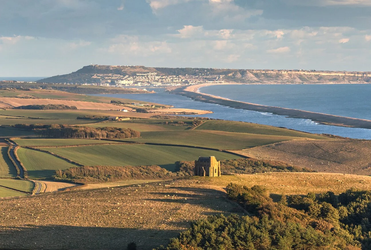 A scenic landscape featuring rolling hills, a coastline with a beach, and a small historic stone building in the foreground.