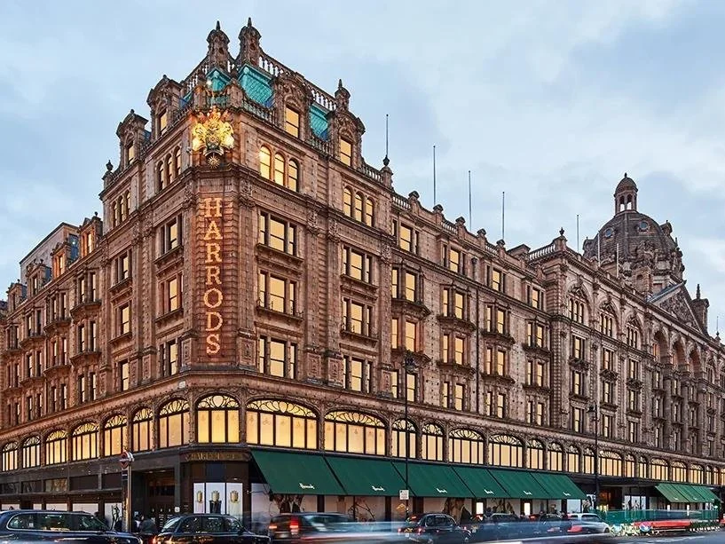 Historic multi-story building with large windows, ornate architecture, and green awnings on the ground floor, with cars parked in front and a city sky background.