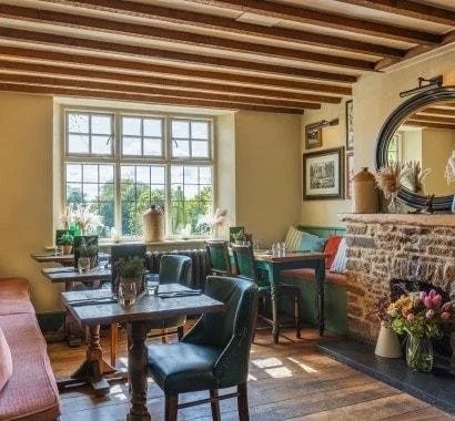 Cozy dining area with wooden ceiling beams, large window, brick fireplace, and decorated with flowers and framed pictures.