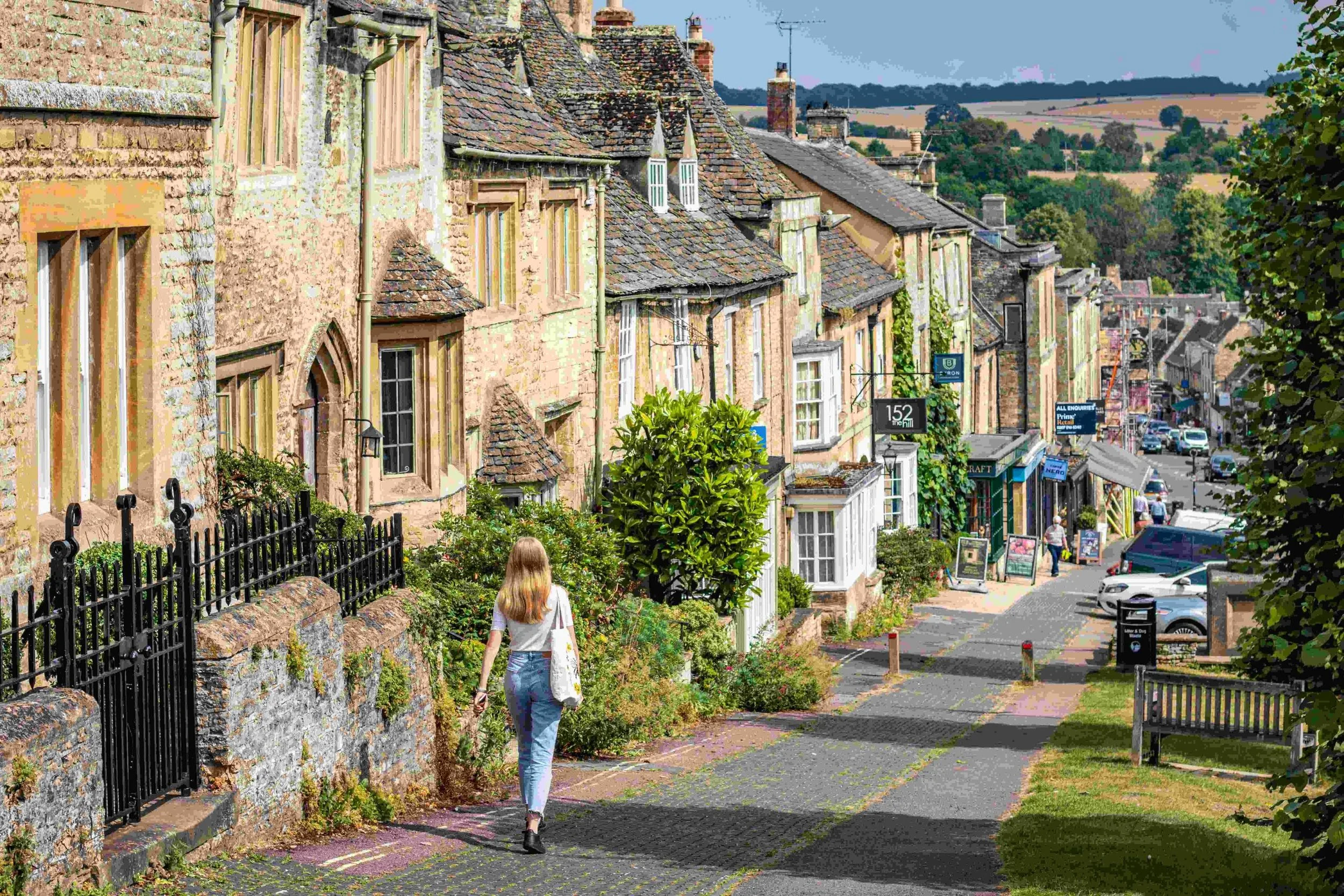 A woman with long hair wearing jeans and a white top, walking on a sidewalk along a street lined with old stone buildings. The buildings have a mix of wooden and stone facades, tiled roofs, and small gardens with iron fences. There are shops, cars parked along the street, and pedestrians visible further down. The background reveals rolling hills and a clear sky, suggesting a small town or village setting.