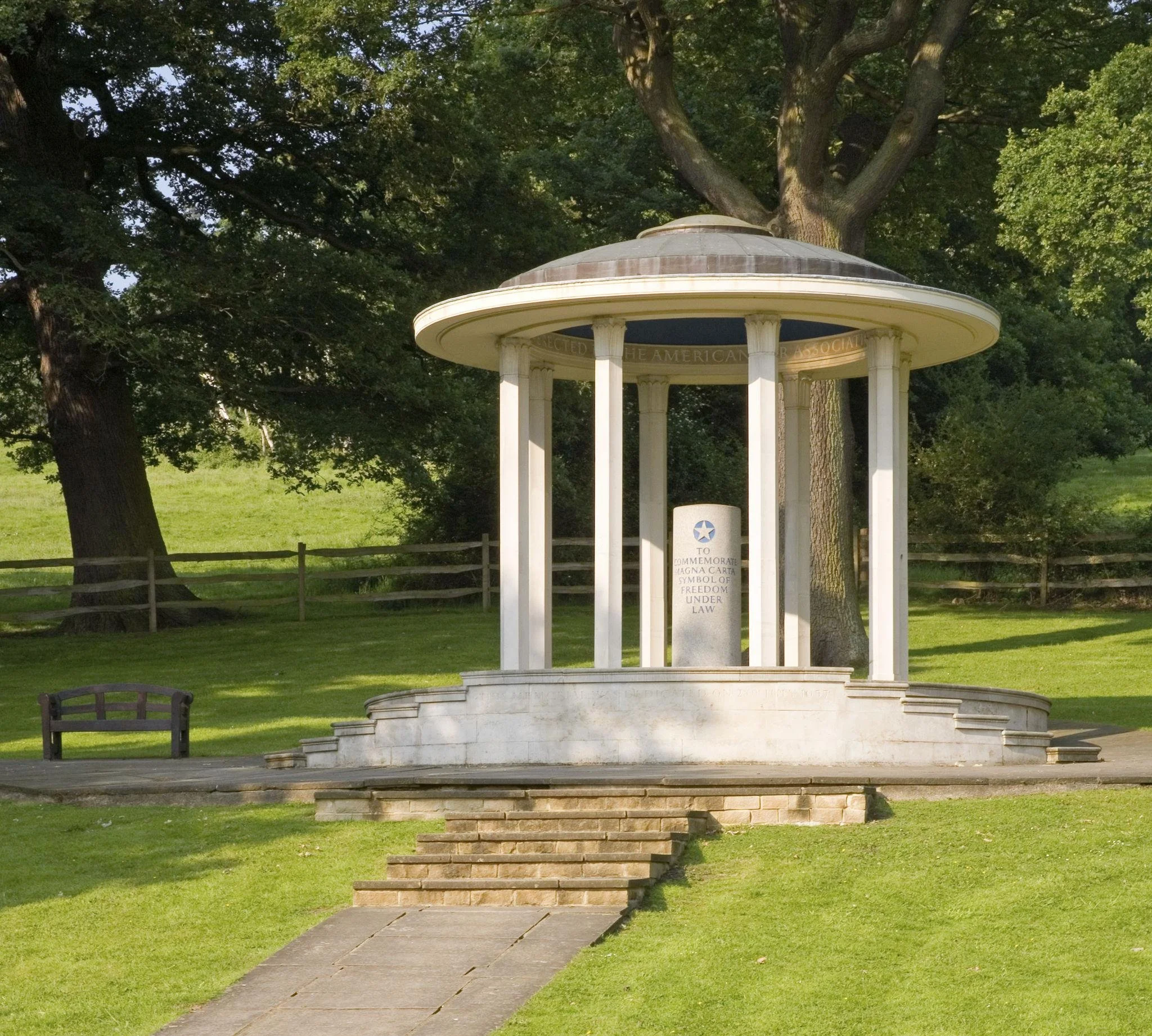 A small white gazebo with a domed roof and columns, situated in a park with green grass and large trees in the background. There's a white stone plaque on the gazebo's base and a wooden fence surrounding the area.
