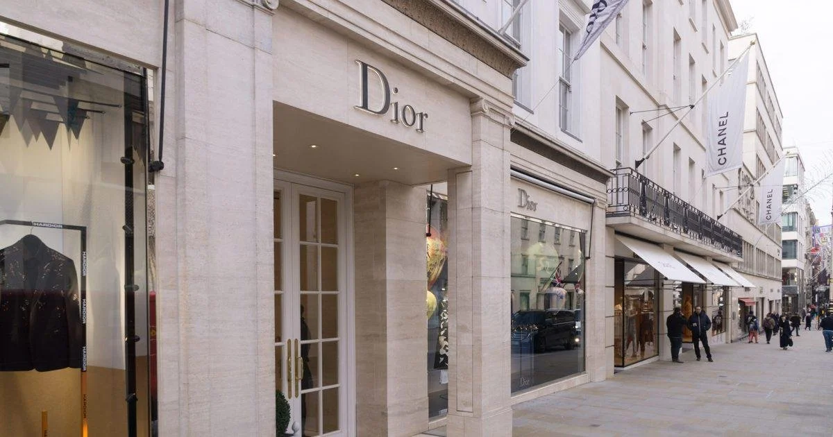 Exterior view of a luxury shopping street with a Dior store in the foreground and a Chanel store further down the street, with shoppers walking by.