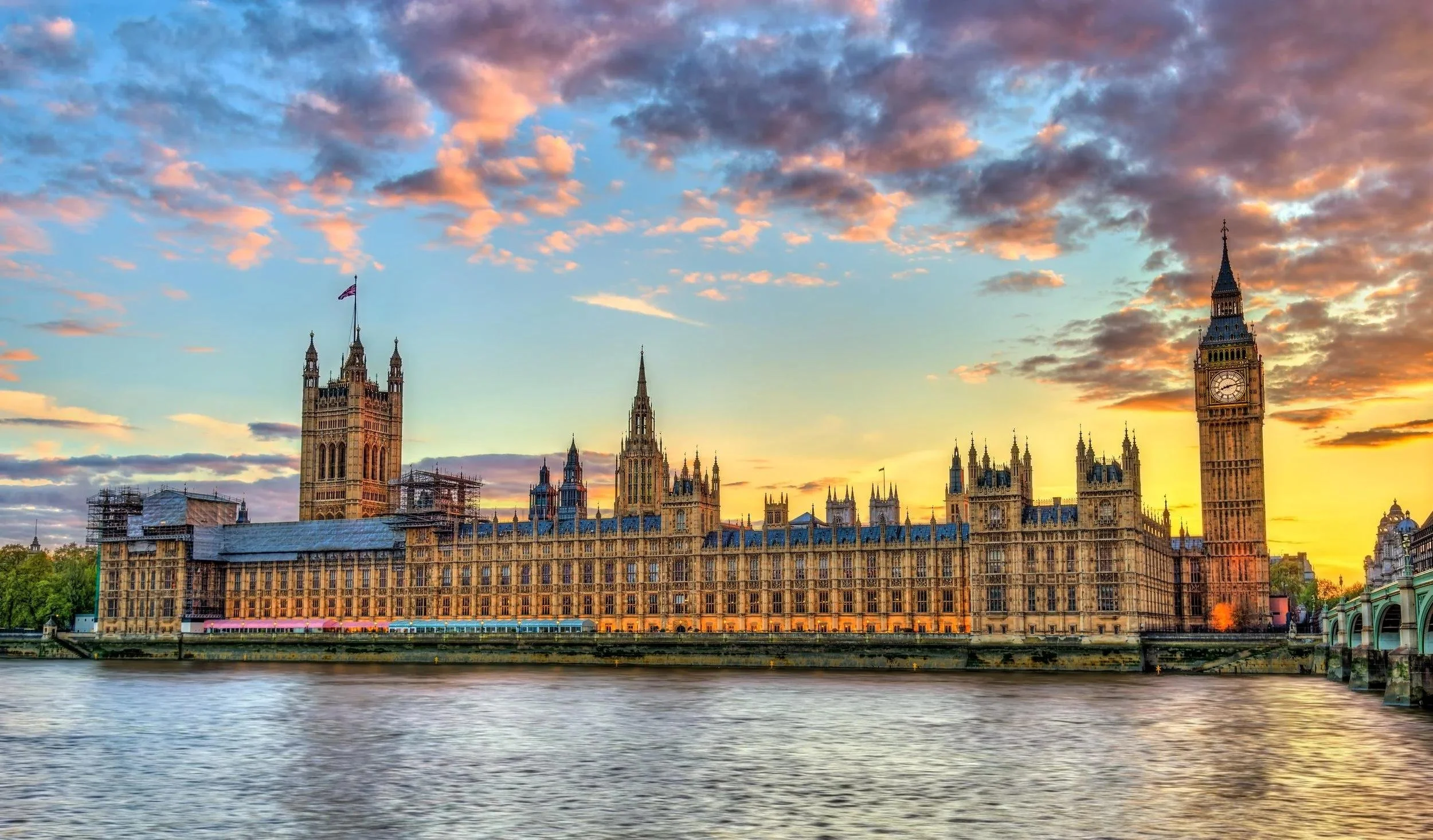 View of the Houses of Parliament in London at sunset with a partly cloudy sky and the River Thames in the foreground.