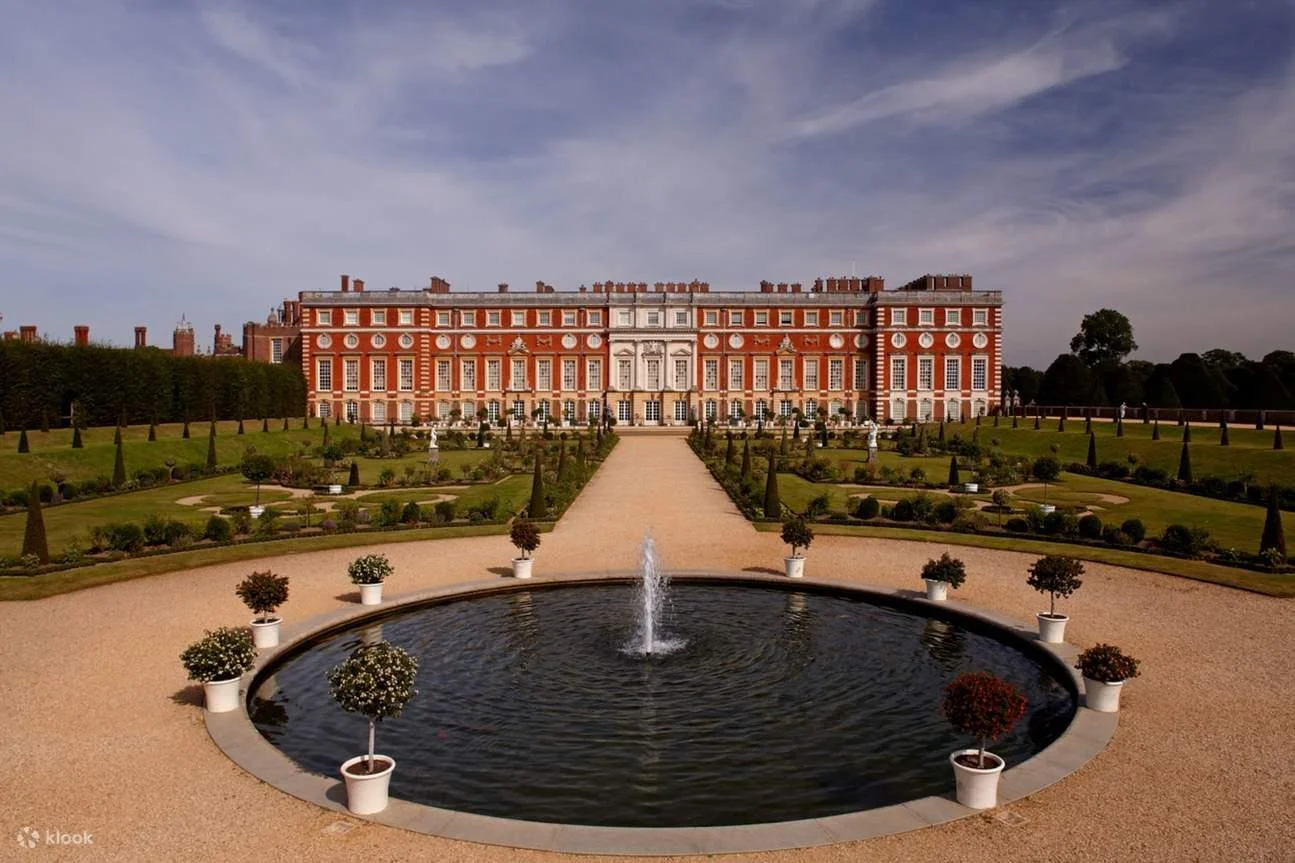 A large historic mansion with red brick walls and white accents, surrounded by a manicured garden with a central fountain and pathway leading up to the building.