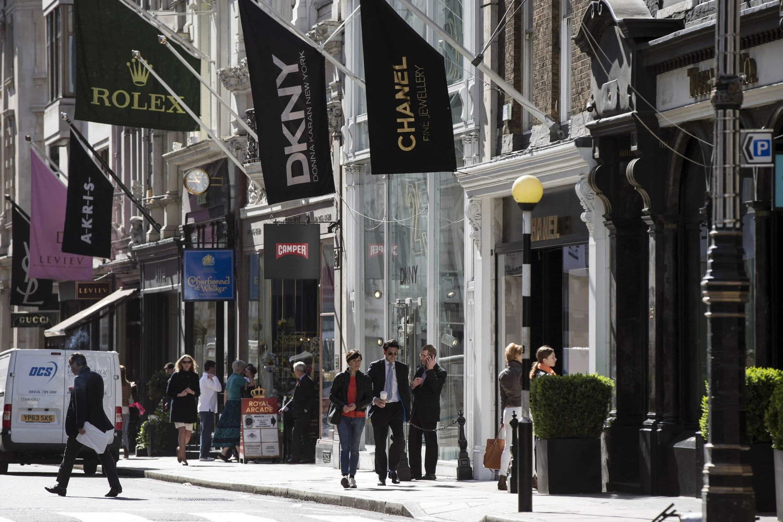 People walking along a busy city street with storefronts, flags, and street lamps.