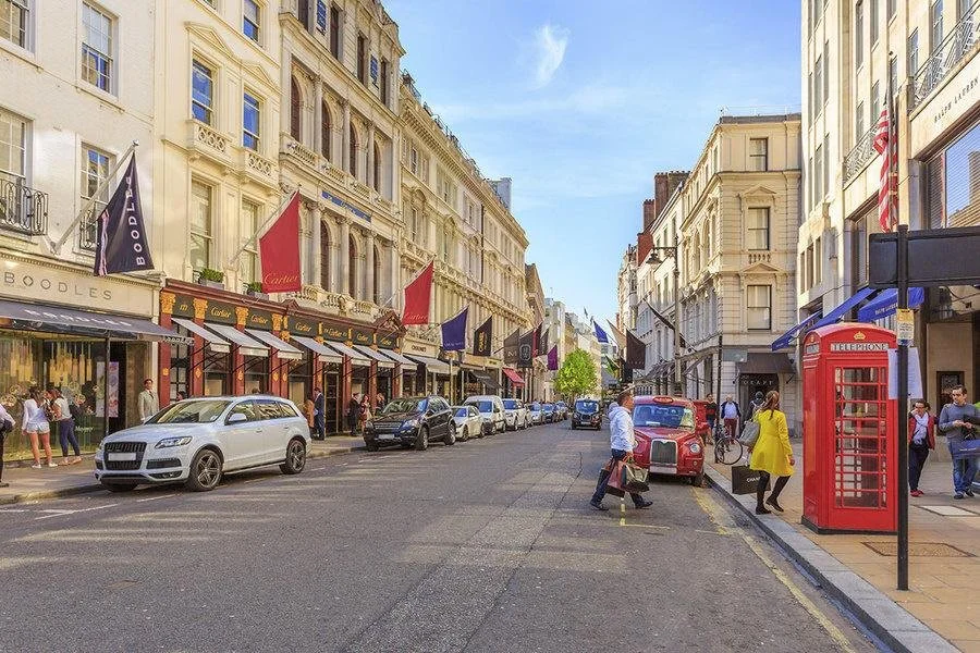 A busy city street with parked cars, pedestrians, and historic buildings. A red telephone booth is visible on the right, with shopping stores on the left, and flags hanging from the buildings.
