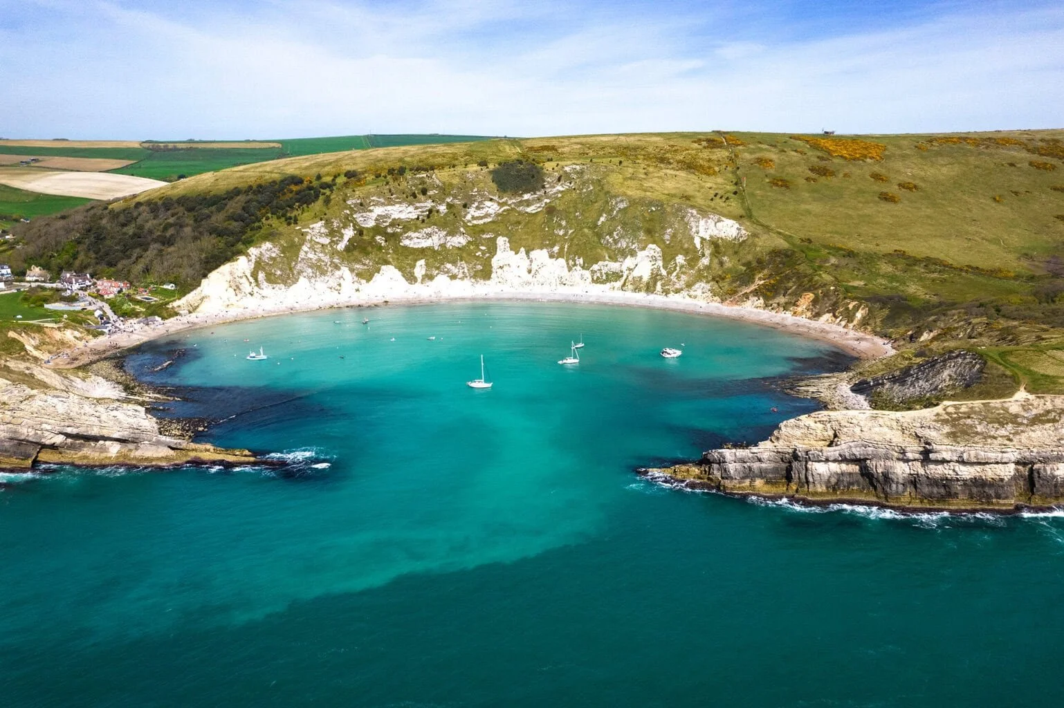A coastal cove with turquoise water, surrounded by green hills and cliffs, and boats anchored in the bay.