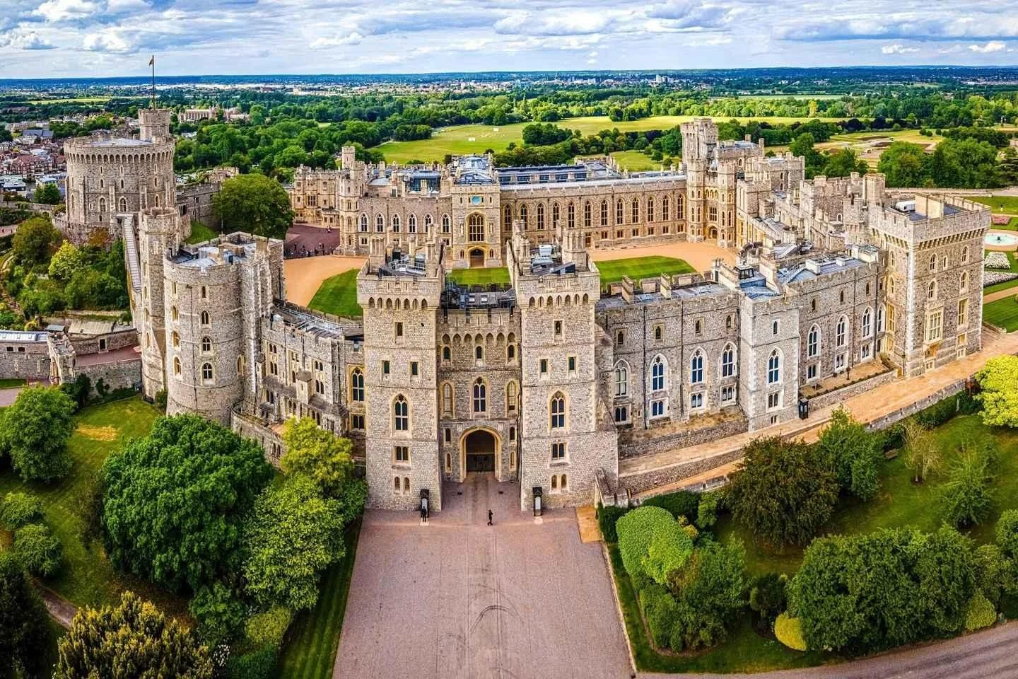 Aerial view of a large, historic stone castle with multiple towers and turrets surrounded by green trees and scenic landscape.