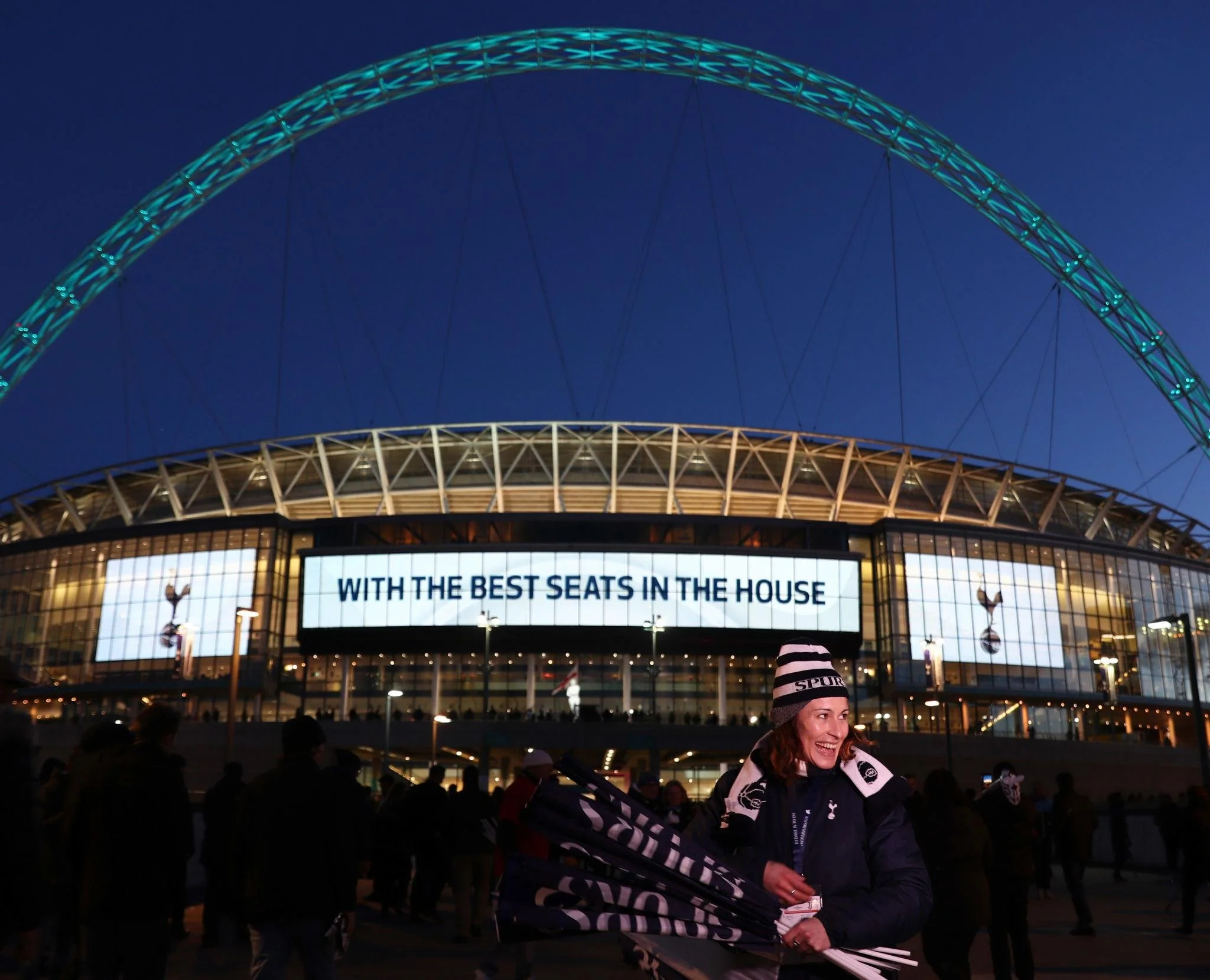 A woman wearing a black and white sports jacket and a beanie hat smiling in front of a large illuminated stadium at night with a large digital sign and a crowd of people around.