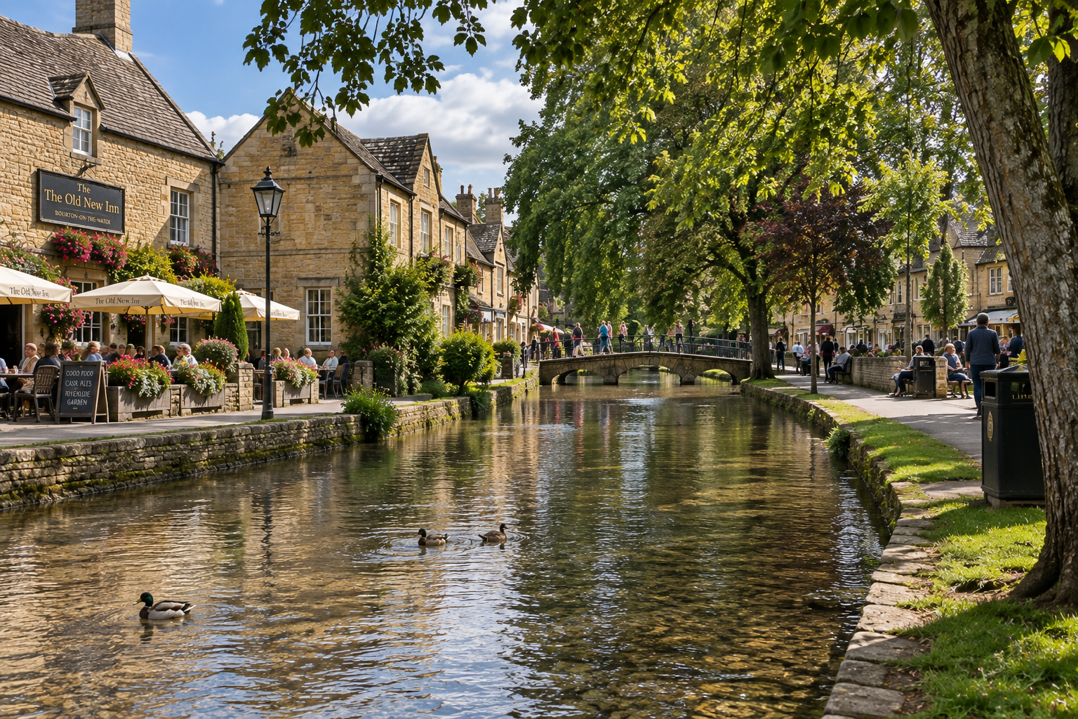 A scenic riverside scene in a small town with historic stone buildings, outdoor dining, a stone bridge, and ducks swimming in the river, on a sunny day with trees and people walking and sitting along the water.