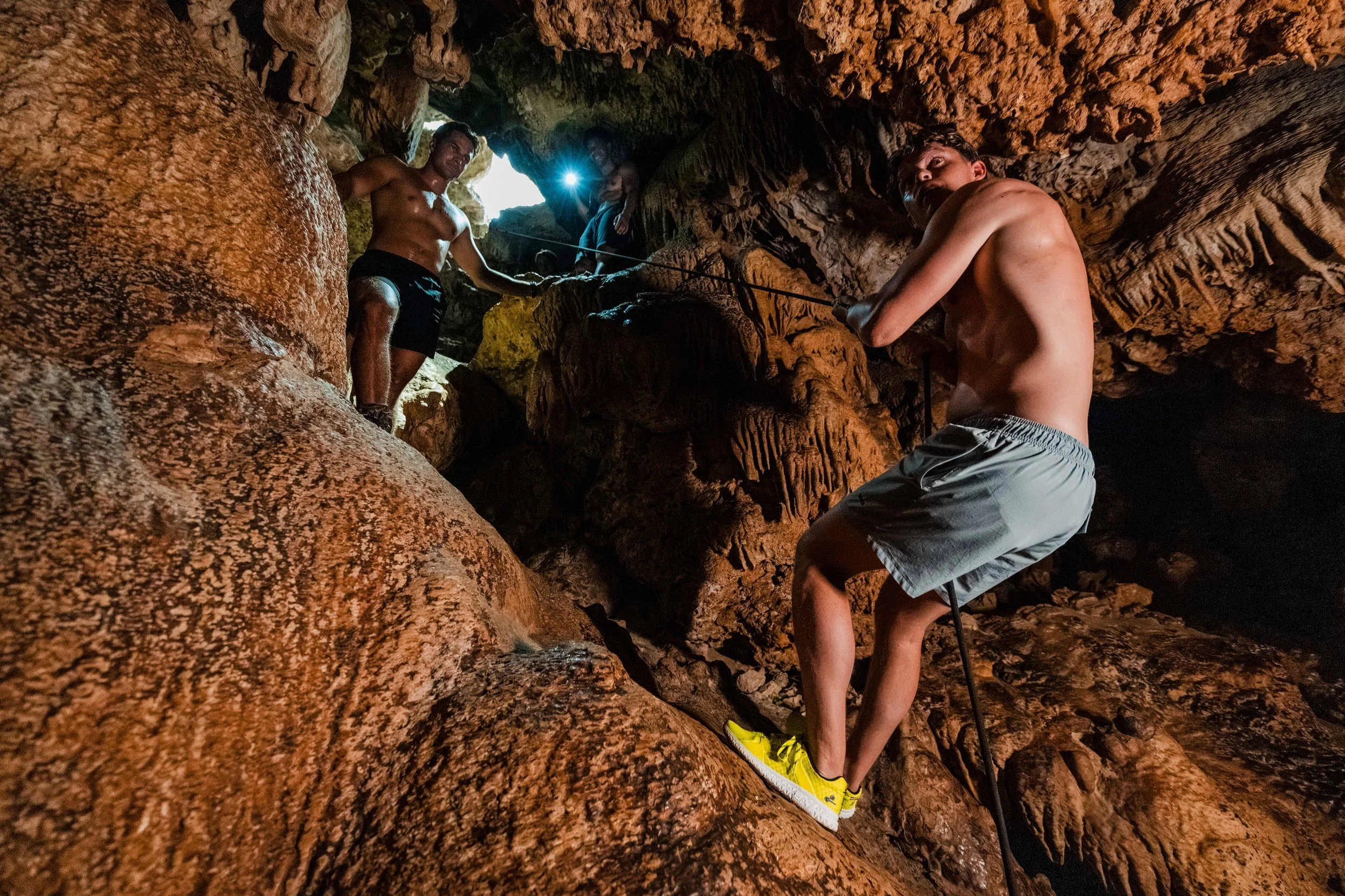 Group of young men exploring a cave, with one man in yellow shoes climbing and others standing nearby. Cave walls are textured with stalactites and stalagmites.