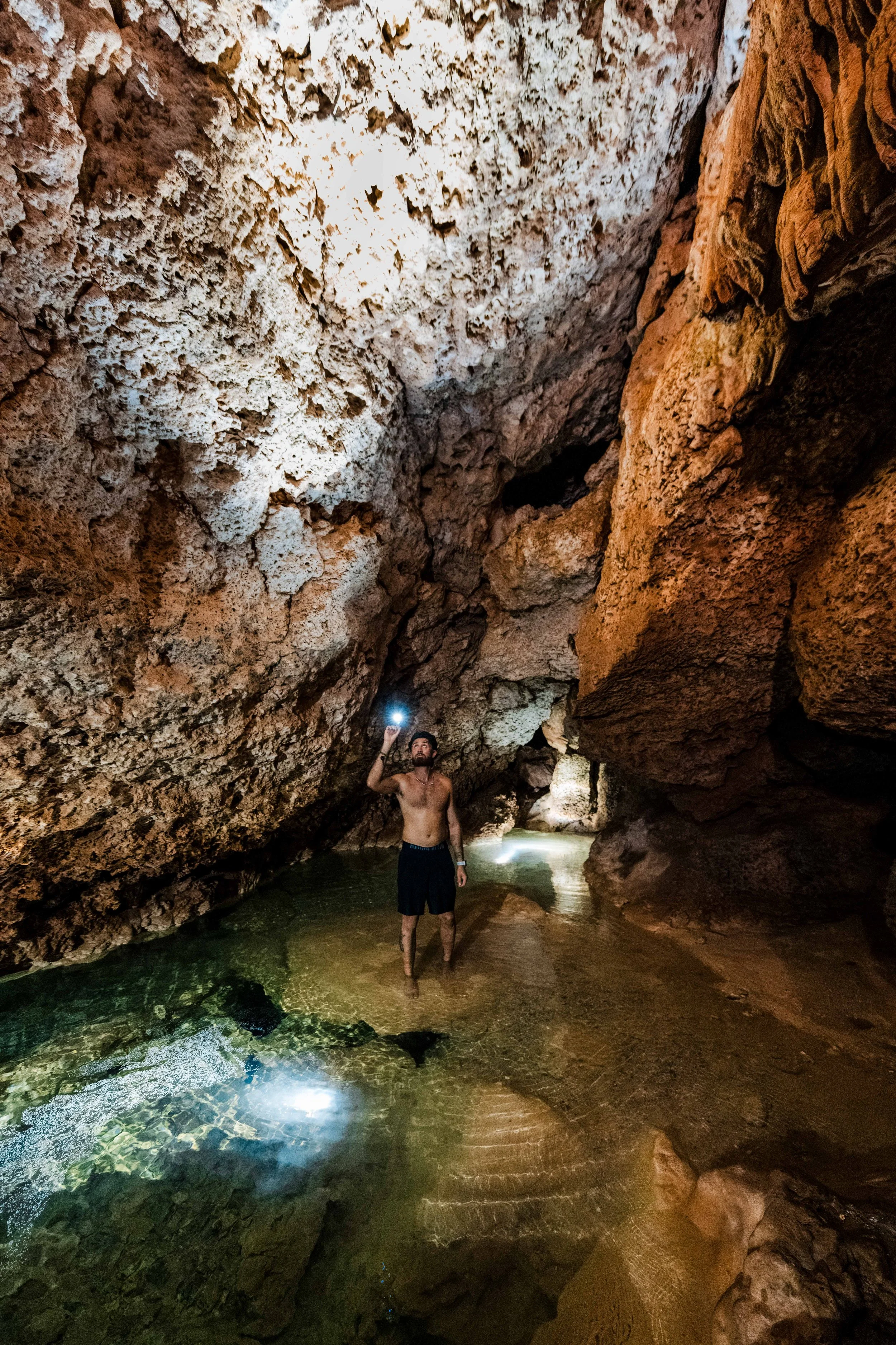 A shirtless man with black shorts standing in a shallow pool of water inside a large cave with tall, textured rock walls and a ceiling. The man is holding a smartphone with the flash on, capturing a photo or video of the cave interior.