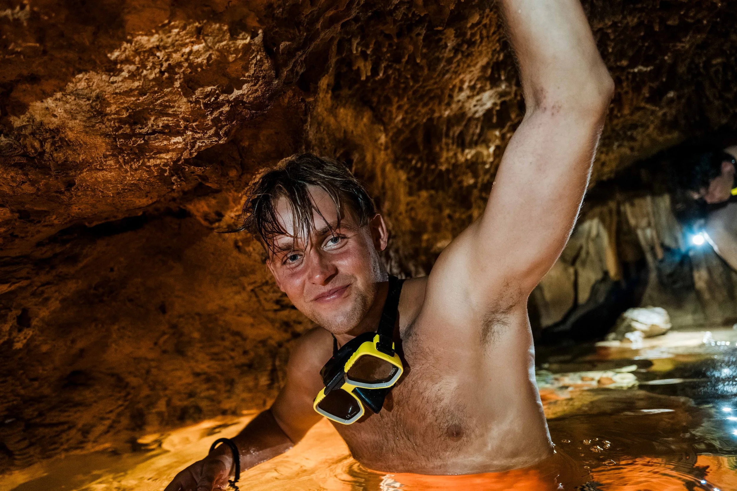 A man with wet hair and a bare chest is climbing or exploring inside a dark underground cave with rocky walls, smiling at the camera.