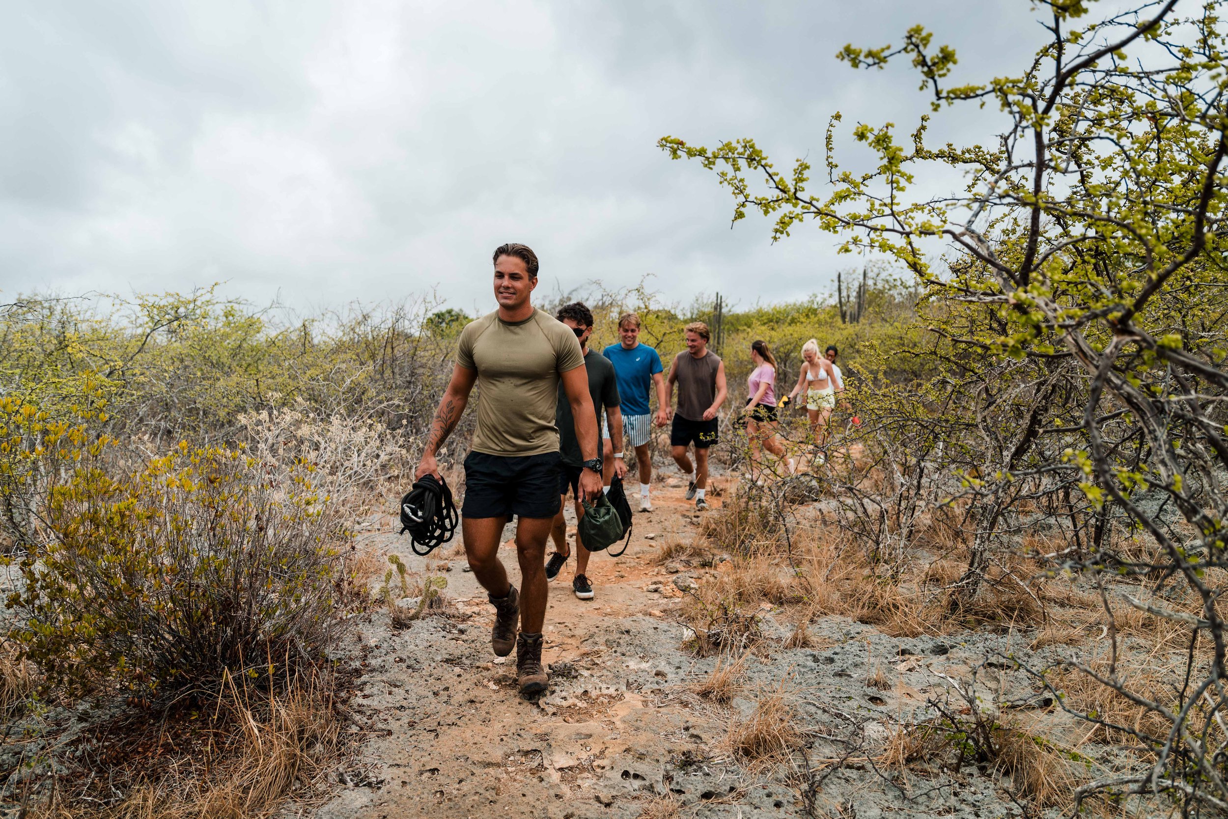 A group of six people walking on a narrow trail through a dry, shrub-covered landscape under an overcast sky. The person at the front is smiling, wearing a tan T-shirt and black shorts, carrying climbing gear.