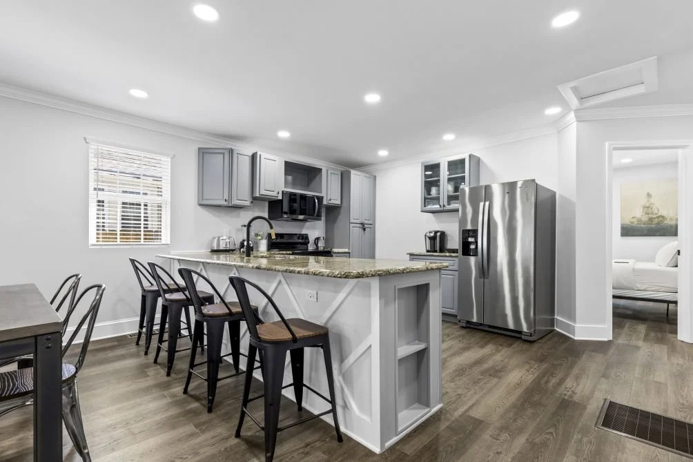 Modern kitchen with gray cabinets, granite countertops, stainless steel refrigerator, microwave, and coffee maker. Barstools at a kitchen island, with a dining table on the left. White walls, wood floors, and a window with blinds.