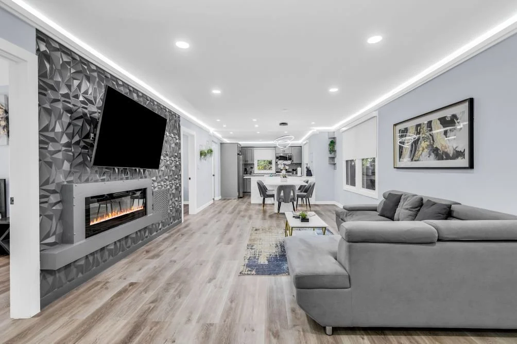 Modern living room with a gray sectional sofa, a wall-mounted TV, and an electric fireplace. There is a dining area and kitchen in the background, with large windows and contemporary decor.