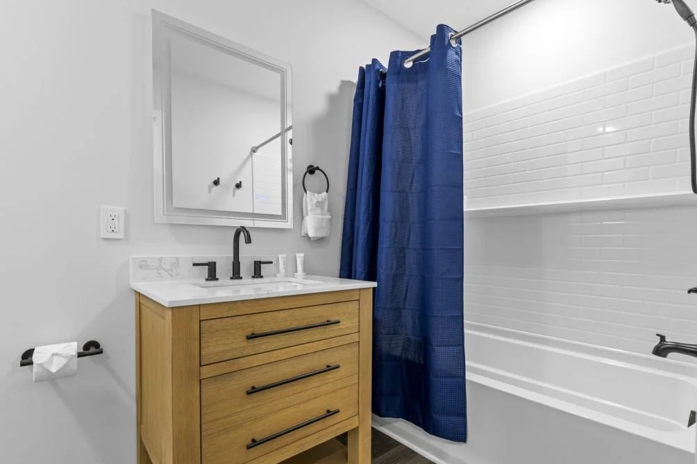 Modern bathroom with white walls, a vanity with a white marble top, black fixtures, a mirror, a toilet paper holder, navy blue shower curtain, and a white bathtub with a built-in shelf.