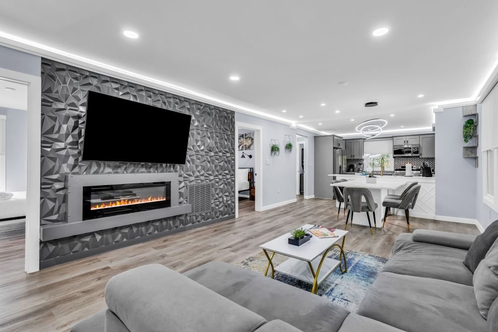 Modern living room with a gray sofa, a white coffee table, a textured gray accent wall with a mounted flat-screen TV and electric fireplace, and an open kitchen with a white island and gray chairs. Recessed lighting and wall-mounted plants adorn the space.