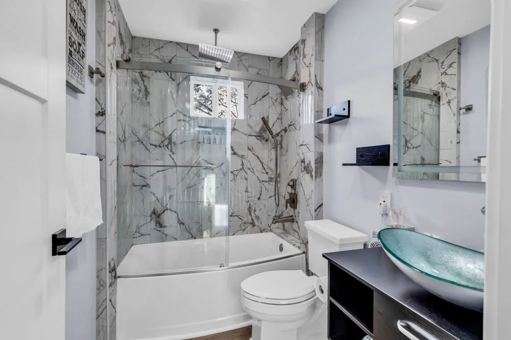 Modern bathroom with marble walls, glass shower with rainfall showerhead, white bathtub, toilet, black vanity with glass vessel sink, mirror, and wall-mounted shelves.