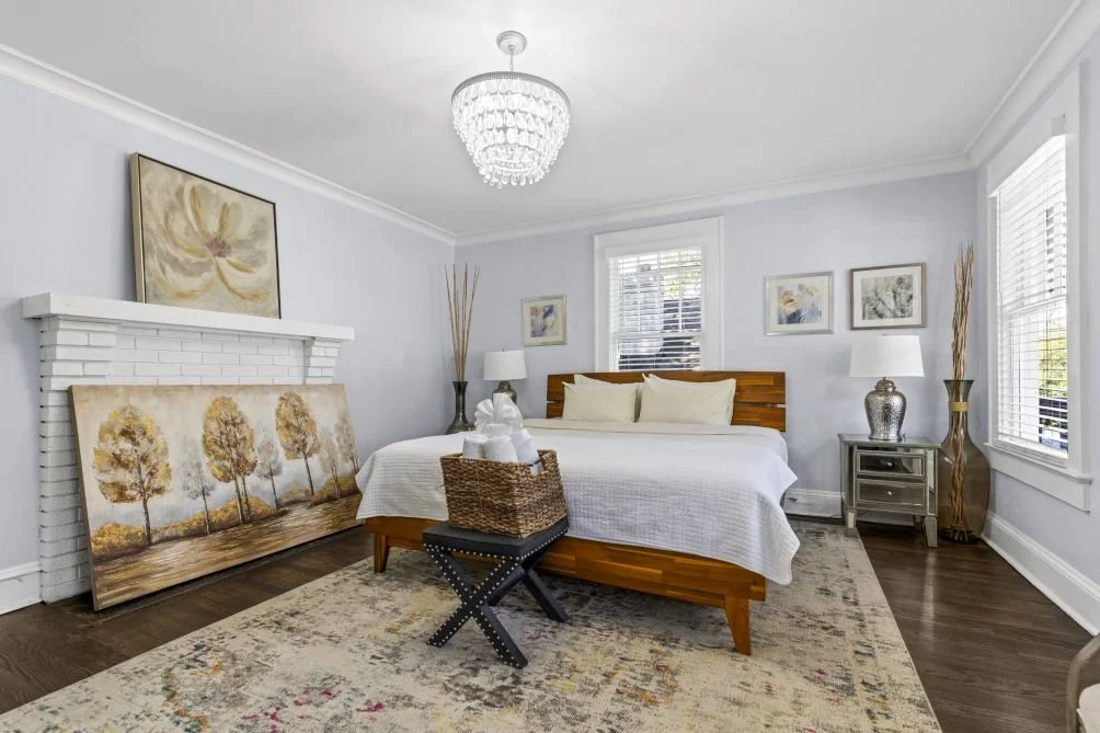 A bedroom with a bed, nightstands with lamps, art on the walls, large windows with blinds, a rug, and a chandelier, decorated in neutral tones.