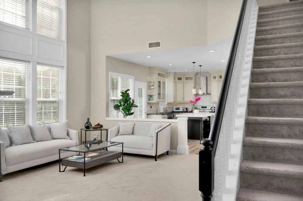 Living room with white sofas, a glass coffee table, and large windows, adjacent to a kitchen with white cabinets and pink flowers on the counter.