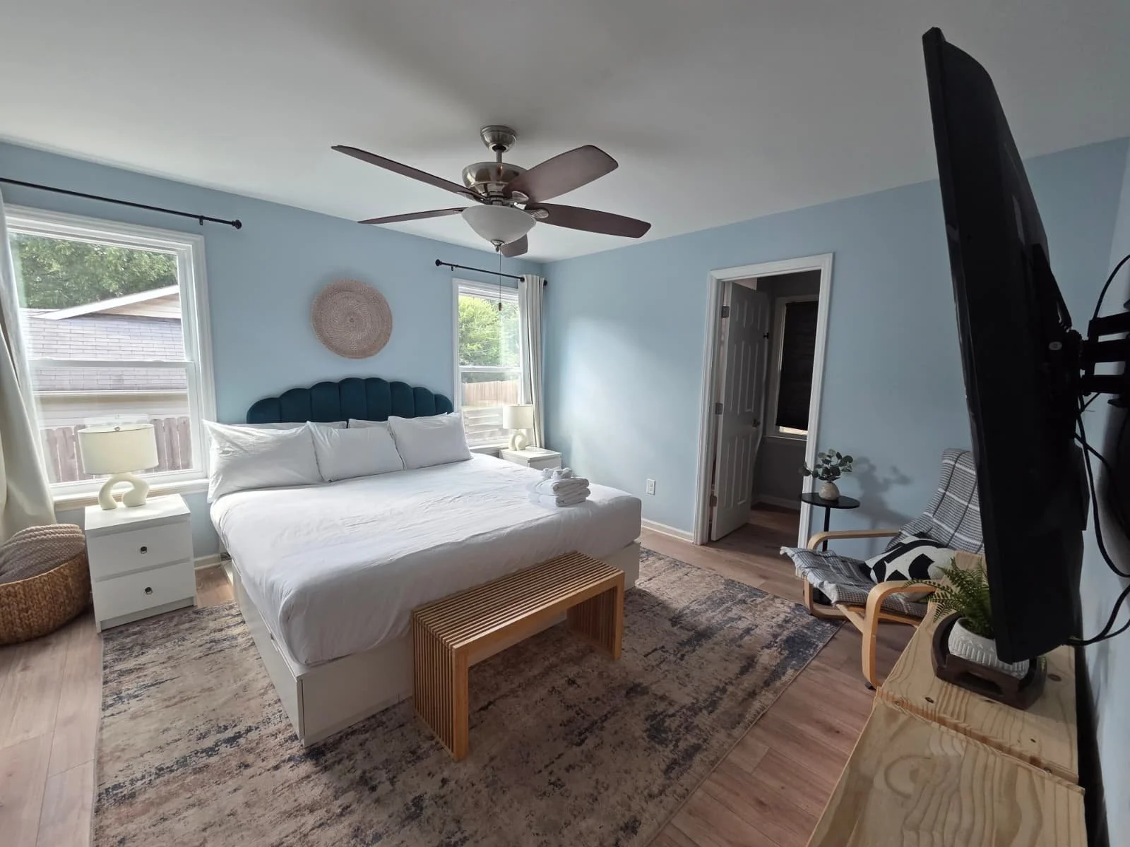 A bedroom with light blue walls, a ceiling fan, two windows with white curtains, a bed with a teal headboard, and white linens. There are two nightstands with lamps, a wicker basket, a wooden bench at the foot of the bed, a TV on a stand, and a chair with plaid upholstery next to a small round table with a plant.