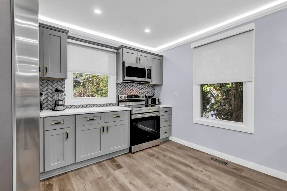 Modern kitchen with gray cabinets, stainless steel appliances, a black and white backsplash, two windows with white blinds, and light wooden flooring.