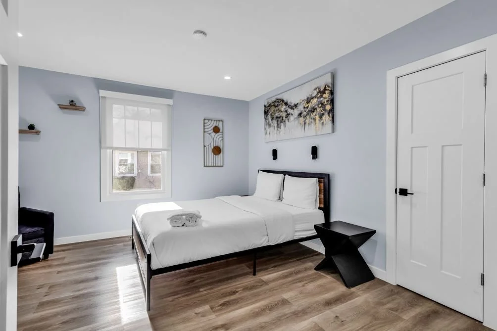 A modern bedroom with light blue walls, a large window, a white bed with two pillows, a black nightstand, black wall-mounted lights, and abstract wall art.